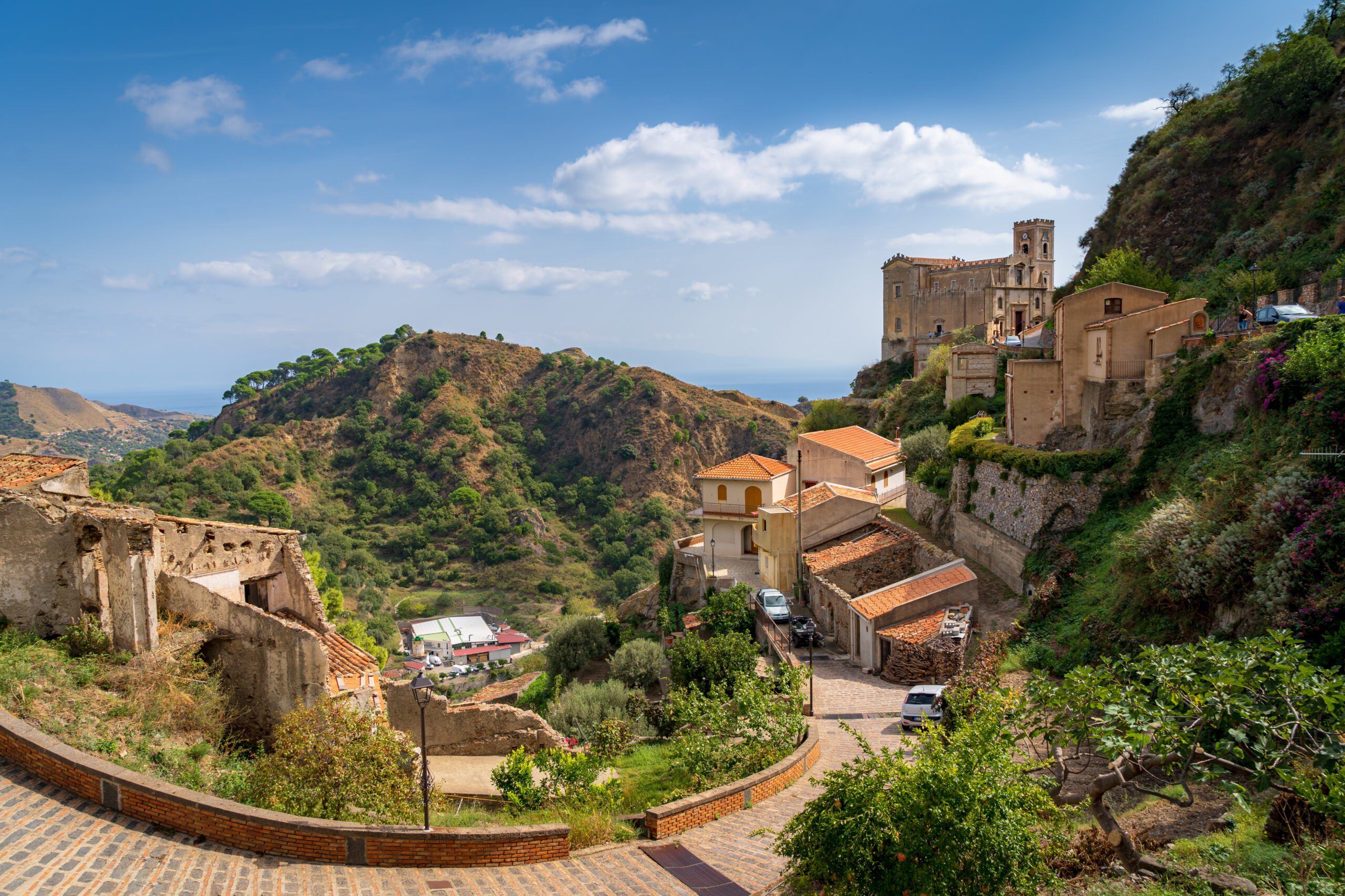 Savoca – kamienne domy i kościół na stromym zboczu, wąska droga prowadząca w dół oraz zielone wzgórza w tle, z daleką panoramą wybrzeża.