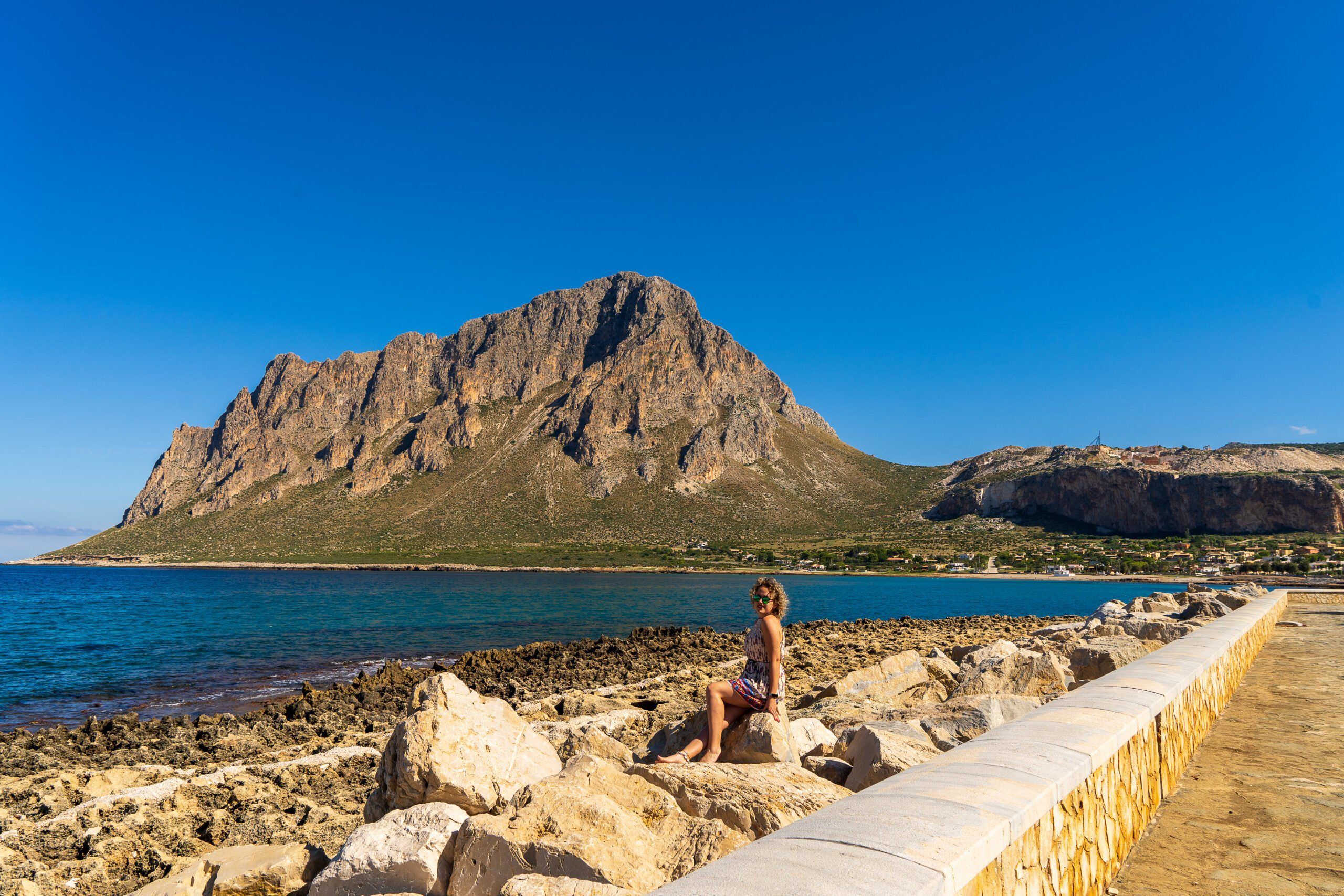 San Vito Lo Capo – kobieta siedząca na skałach przy nadmorskim deptaku z widokiem na majestatyczną górę Monte Monaco