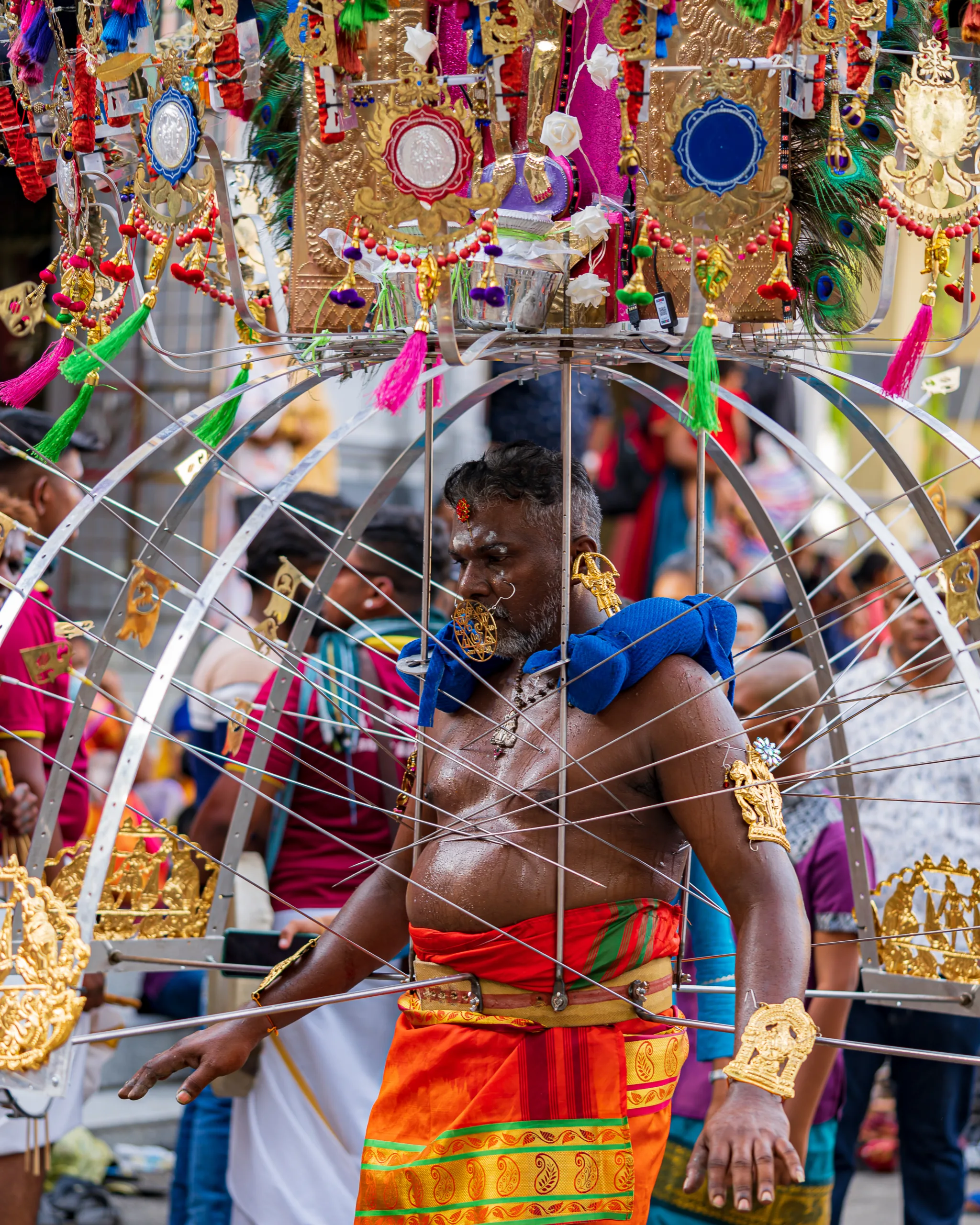 Mężczyzna biorący udział w procesji Thaipusam w Malezji, ubrany w tradycyjny strój, z ozdobami i metalową konstrukcją przebijającą ciało.