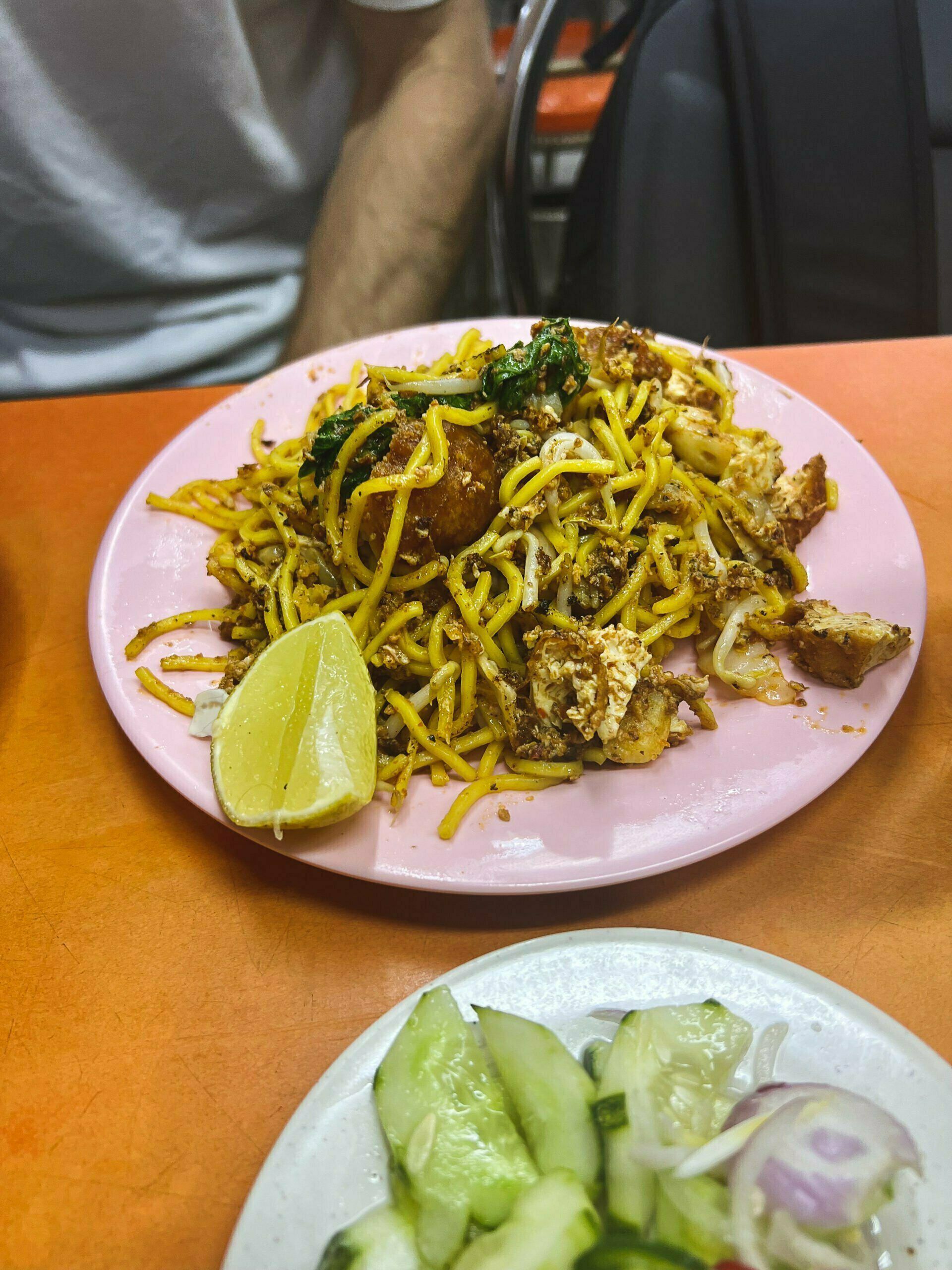 Talerz smażonego makaronu z tofu, jajkiem, warzywami i kawałkiem limonki, podany na różowym talerzu, obok sałatka z ogórka i cebuli - Mee goreng - Kuchnia Malezyjska