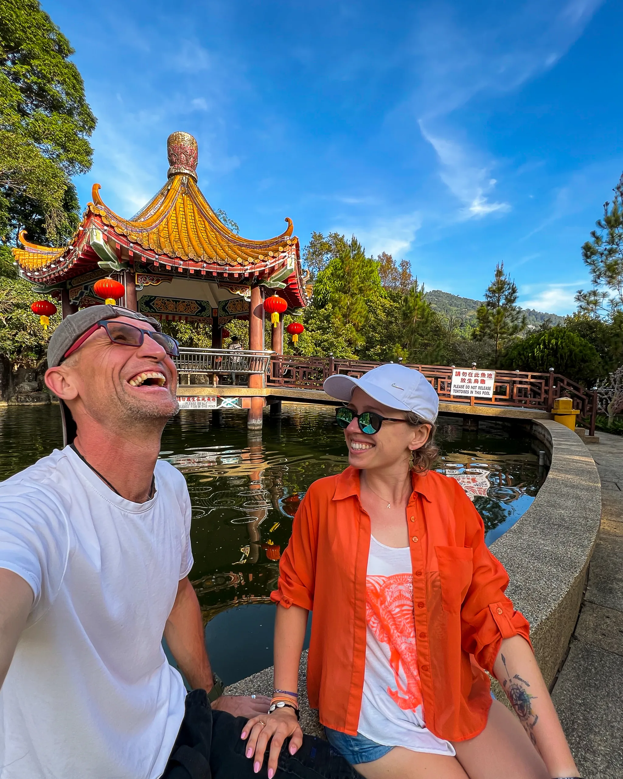 Para uśmiechająca się przed tradycyjną chińską altaną nad stawem, w słoneczny dzień.