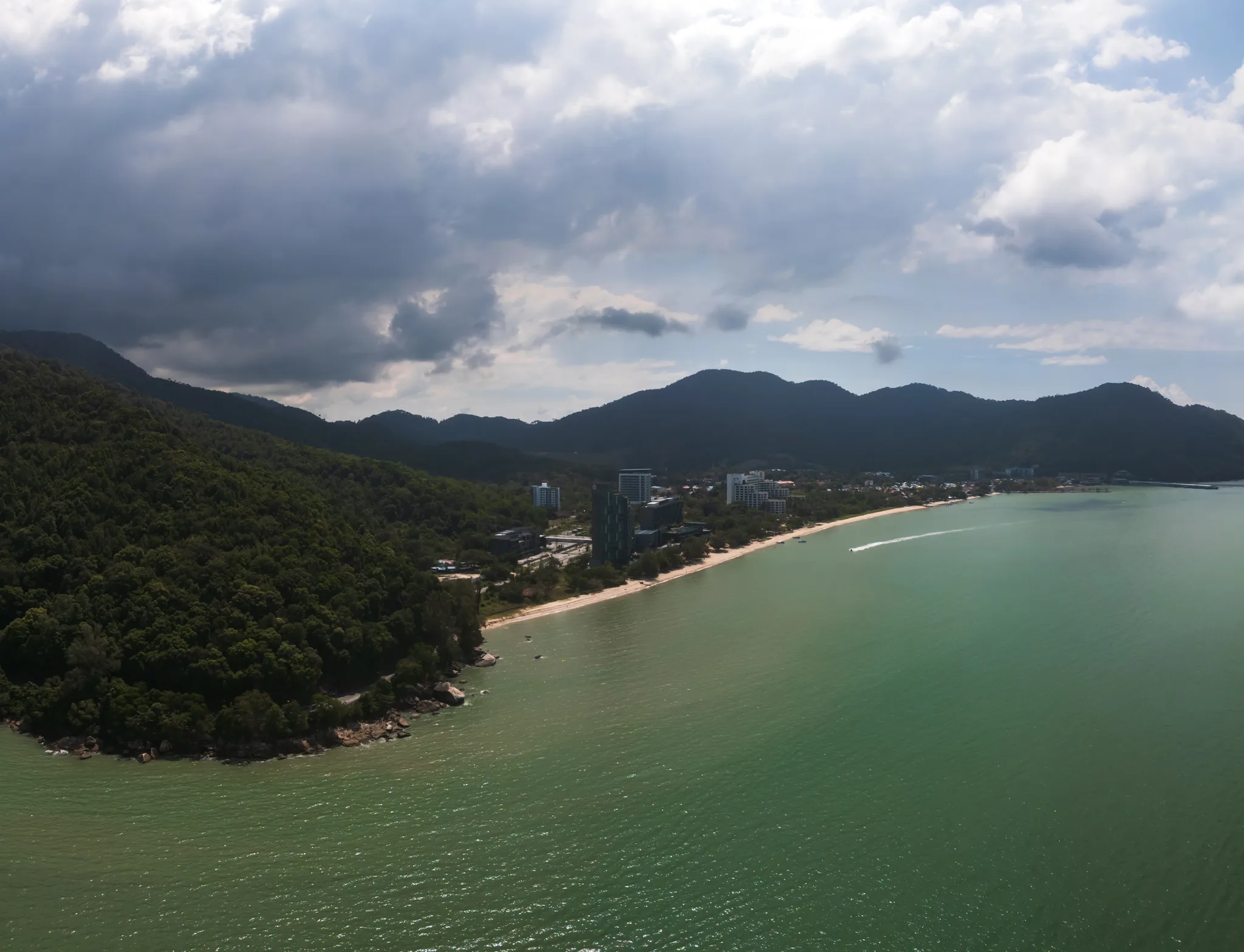 Panorama wybrzeża Penang – zielone wzgórza schodzące do morza, złota plaża, turkusowa woda oraz nadciągające ciemne chmury burzowe.