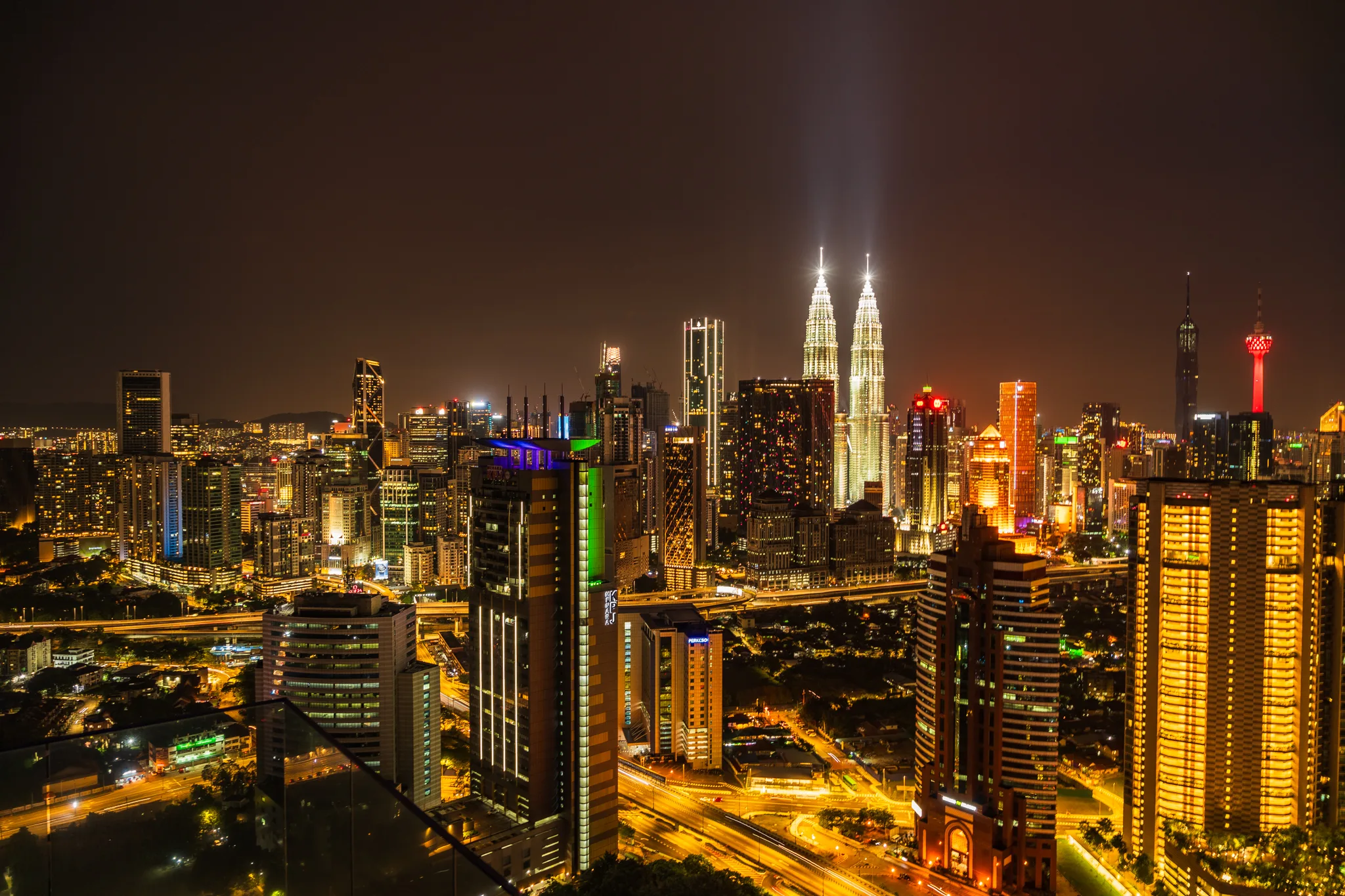 Panorama nocnego Kuala Lumpur z widokiem na oświetlone wieże Petronas Twin Towers oraz inne drapacze chmur.