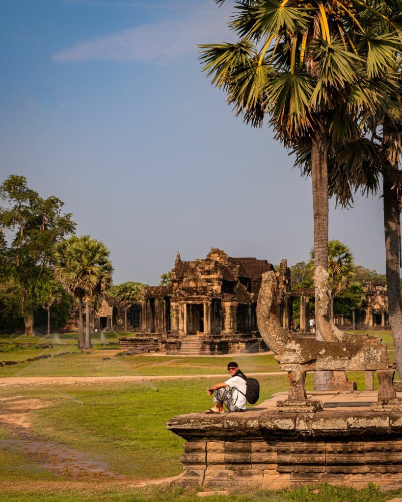 Podróżnik siedzący wśród starożytnych ruin Angkor Wat w Kambodży, otoczonych palmami i zielenią.