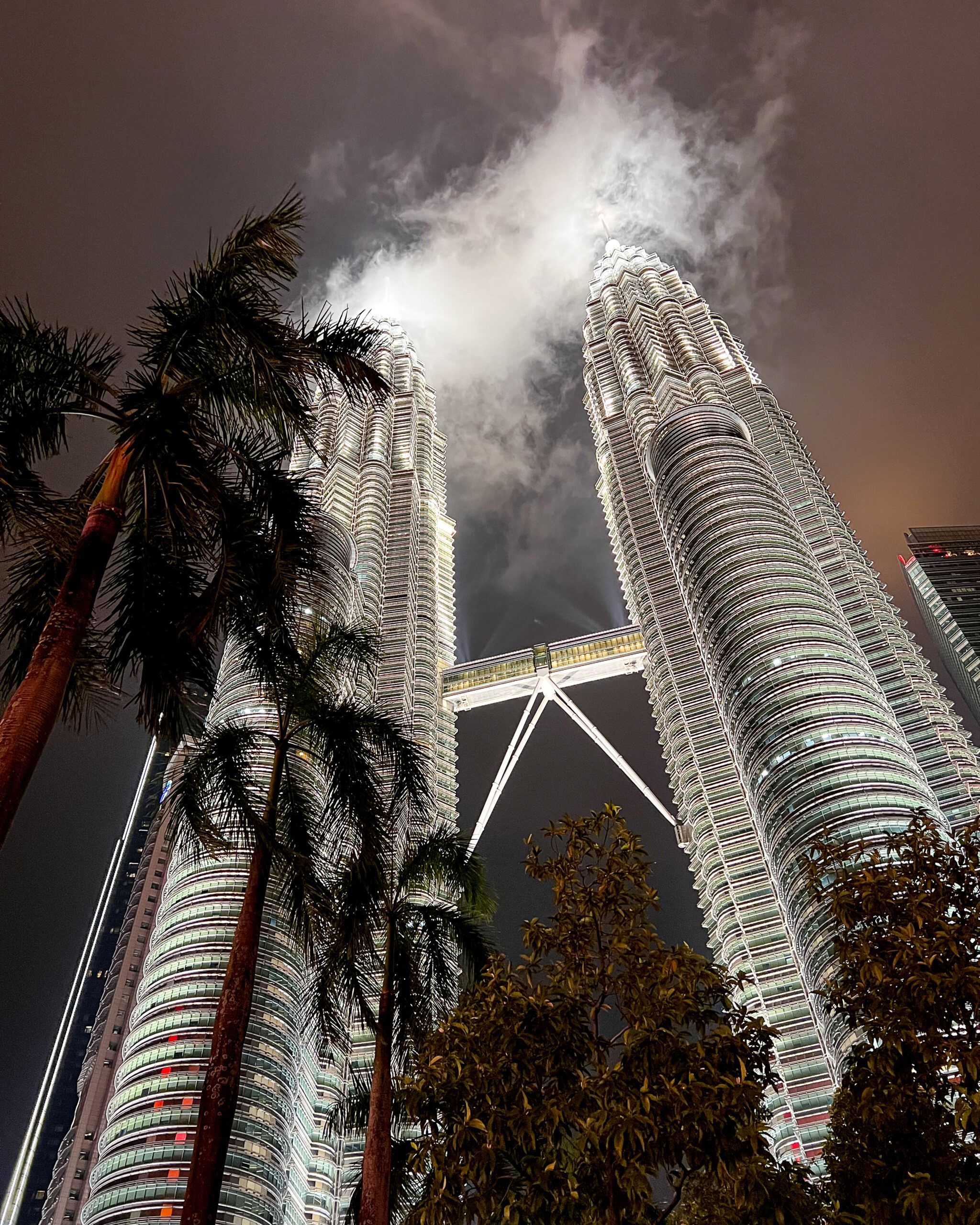 Nocny widok na oświetlone wieże Petronas w Kuala Lumpur z perspektywy od dołu, z palmami i chmurami nad szczytami.