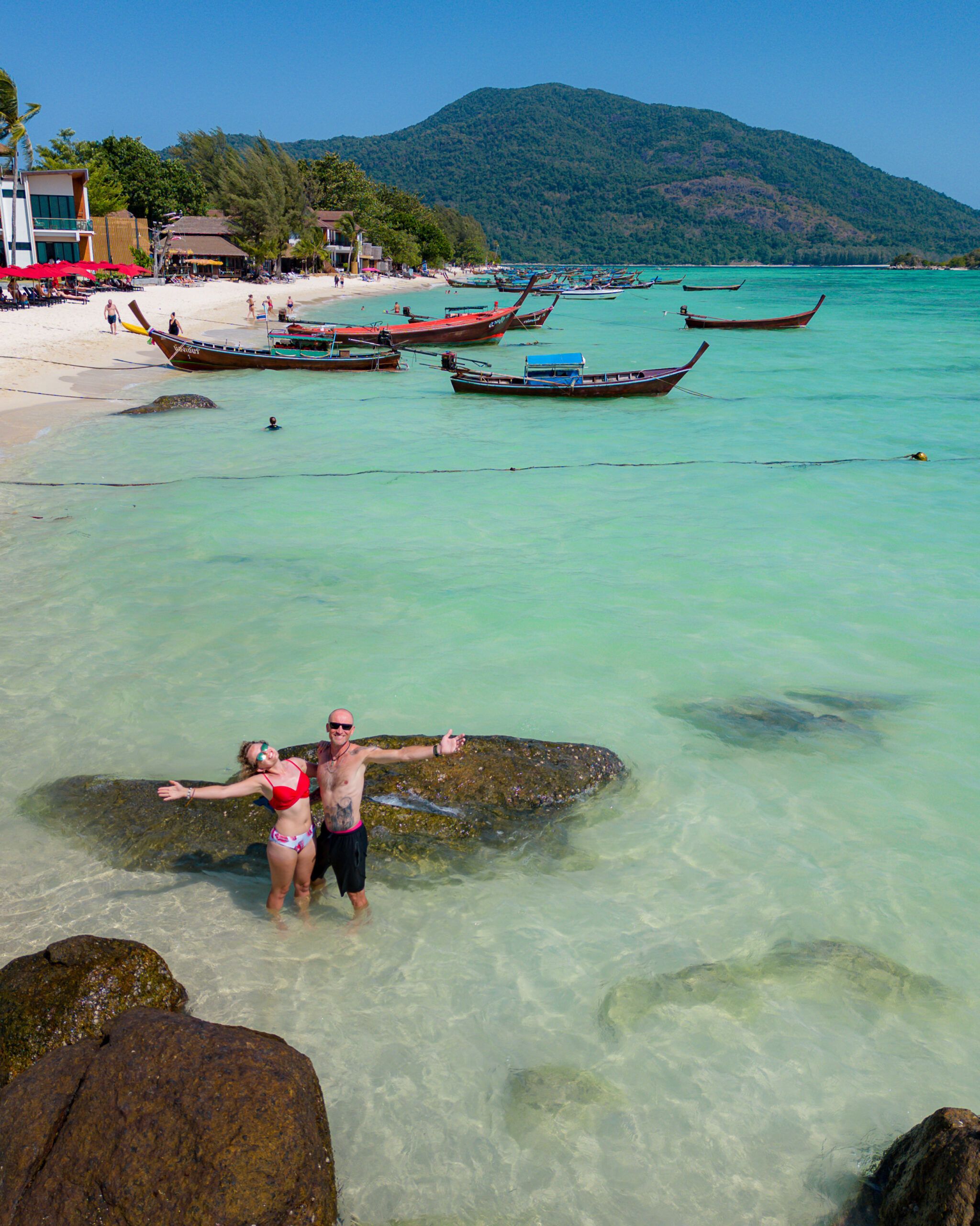 Para stojąca w turkusowej wodzie przy plaży Sunrise Beach na Koh Lipe, w tle tradycyjne łodzie longtail i góry