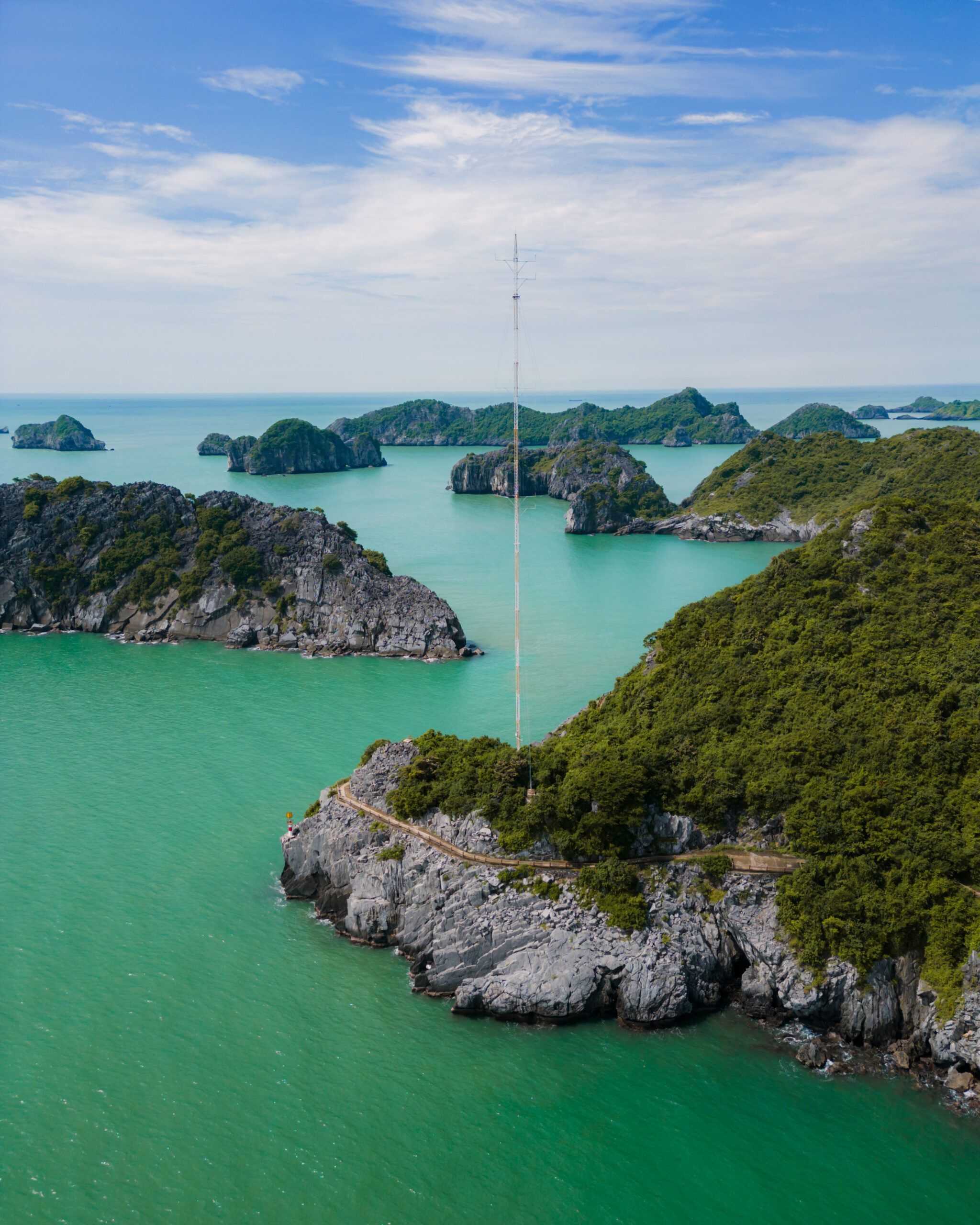 Widok z lotu ptaka na skaliste wybrzeże Cat Ba z wijącą się ścieżką i masztem radiowym, otoczone lazurową wodą i wyspami zatoki Lan Ha