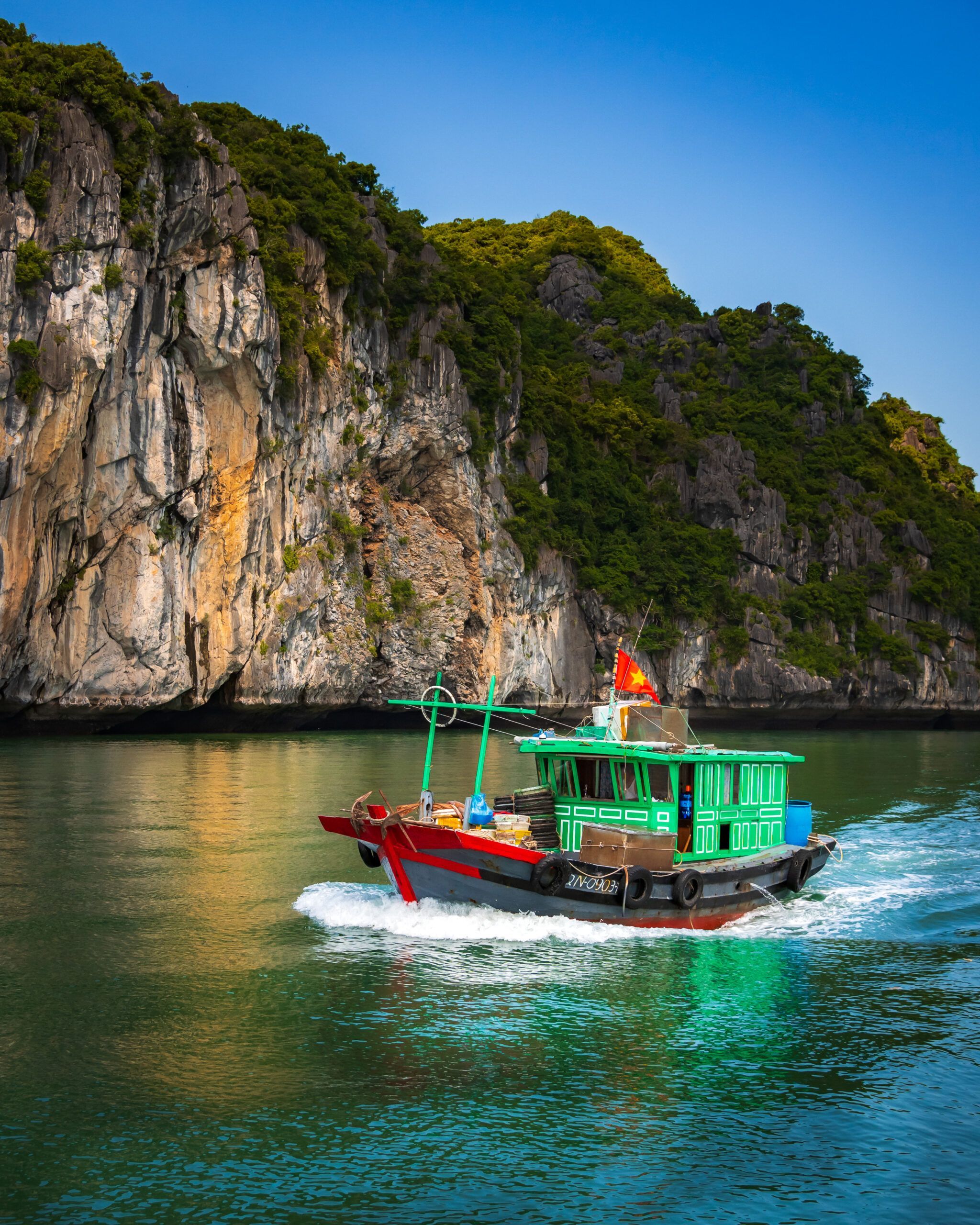 Kolorowa wietnamska łódź rybacka płynąca po spokojnych wodach zatoki Lan Ha, w tle strome wapienne klify porośnięte zielenią