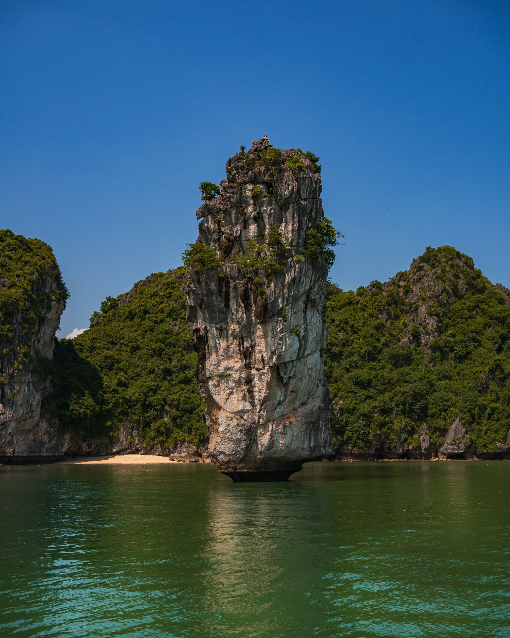 Wysoka wapienna skała wystająca pionowo z wody w zatoce Lan Ha, otoczona zielonymi wyspami i błękitnym niebem