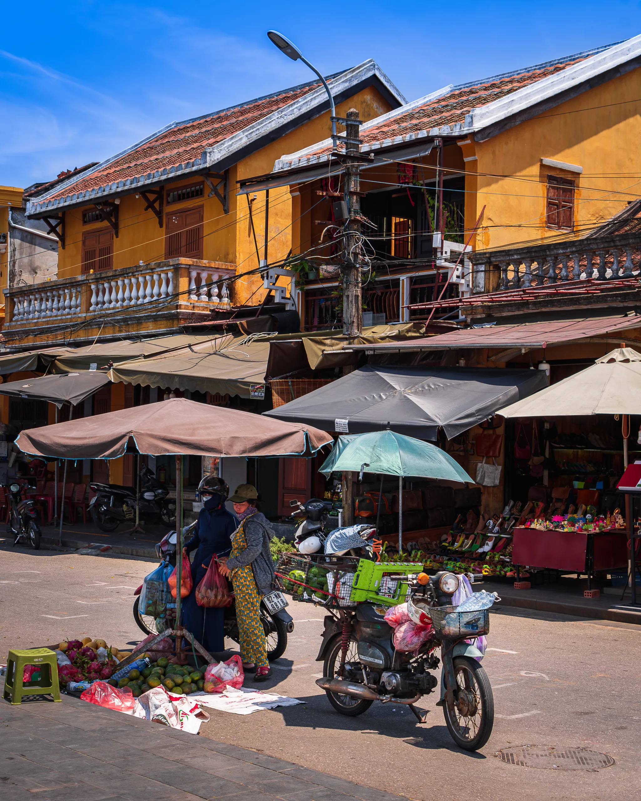 Uliczny targ w Hoi An z kobietami sprzedającymi owoce pod parasolami, motocyklami i kolonialną architekturą w tle