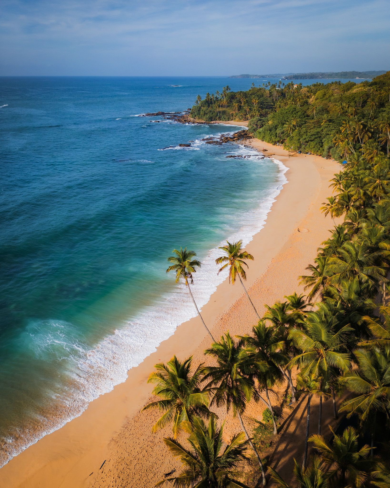 Złocista plaża na Sri Lance otoczona palmami kokosowymi, z turkusowymi falami oceanu rozbijającymi się o brzeg.