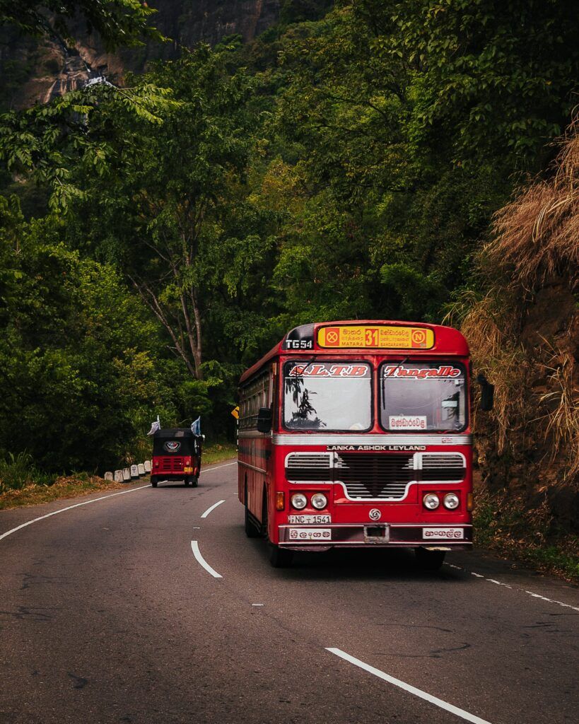 Czerwony lankijski autobus Ashok Leyland jadący krętą drogą przez zielone wzgórza Sri Lanki.
