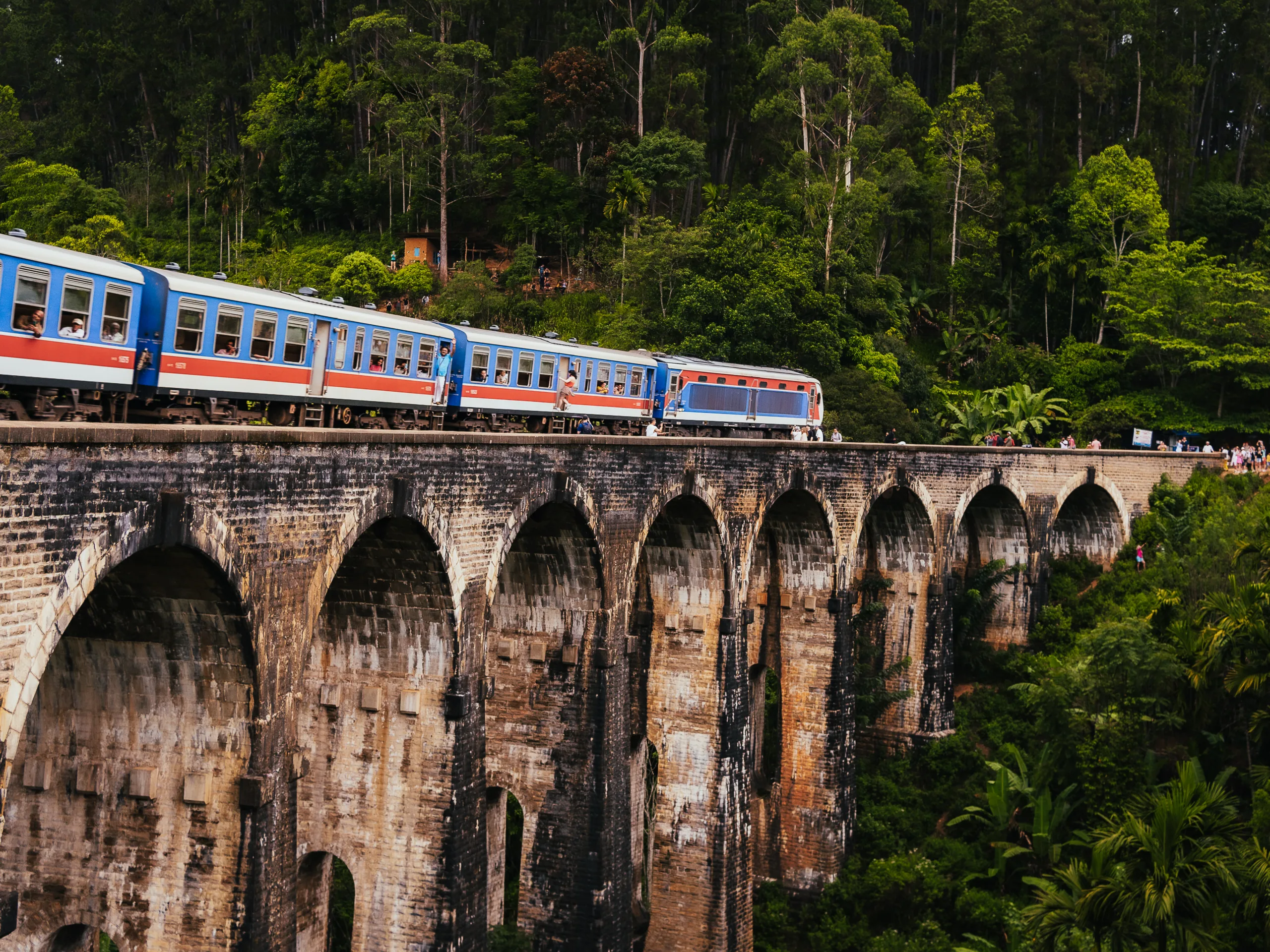 Słynny most dziewięciu łuków na Sri Lance w Ella z przejeżdżającym niebiesko-czerwonym pociągiem otoczonym zieloną dżunglą.