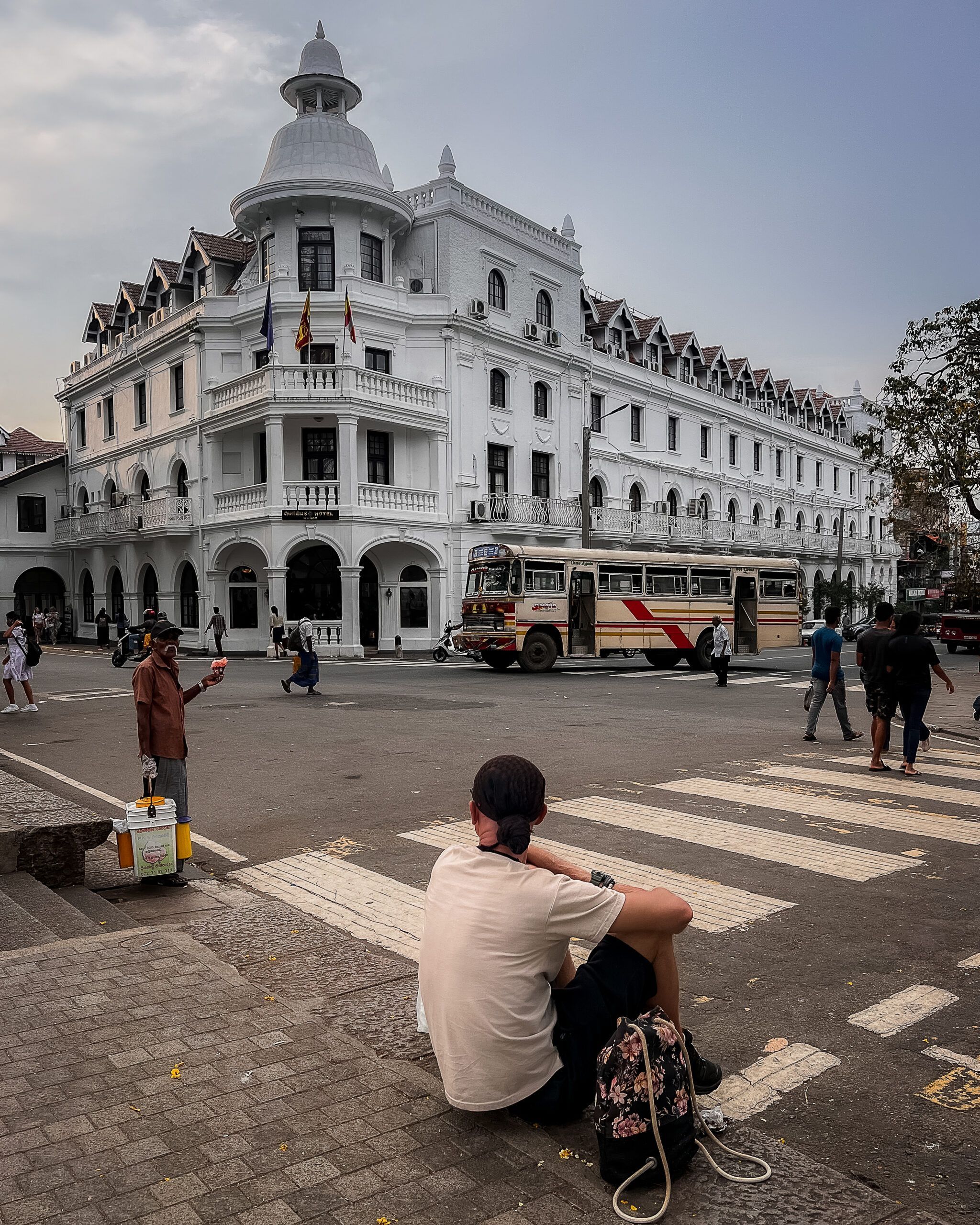Kolonialny biały budynek w centrum Kandy na Sri Lance, przed którym przejeżdża autobus, a turysta siedzi na chodniku obserwując ruch uliczny.