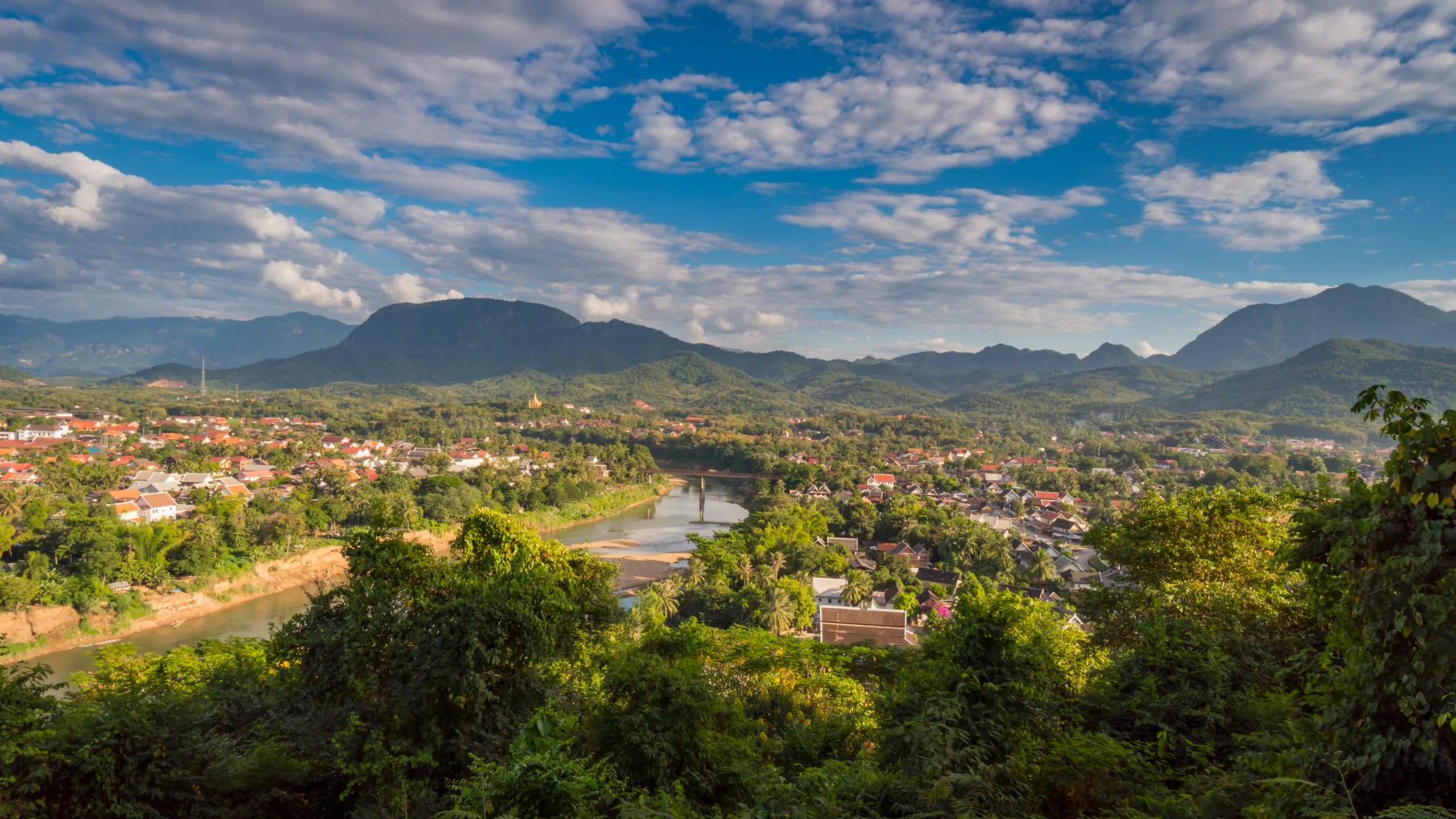 Panorama Luang Prabang w Laosie widziana z góry Phousi, z rzeką, zielonymi wzgórzami i zabudowaniami miasta pod błękitnym niebem z chmurami.