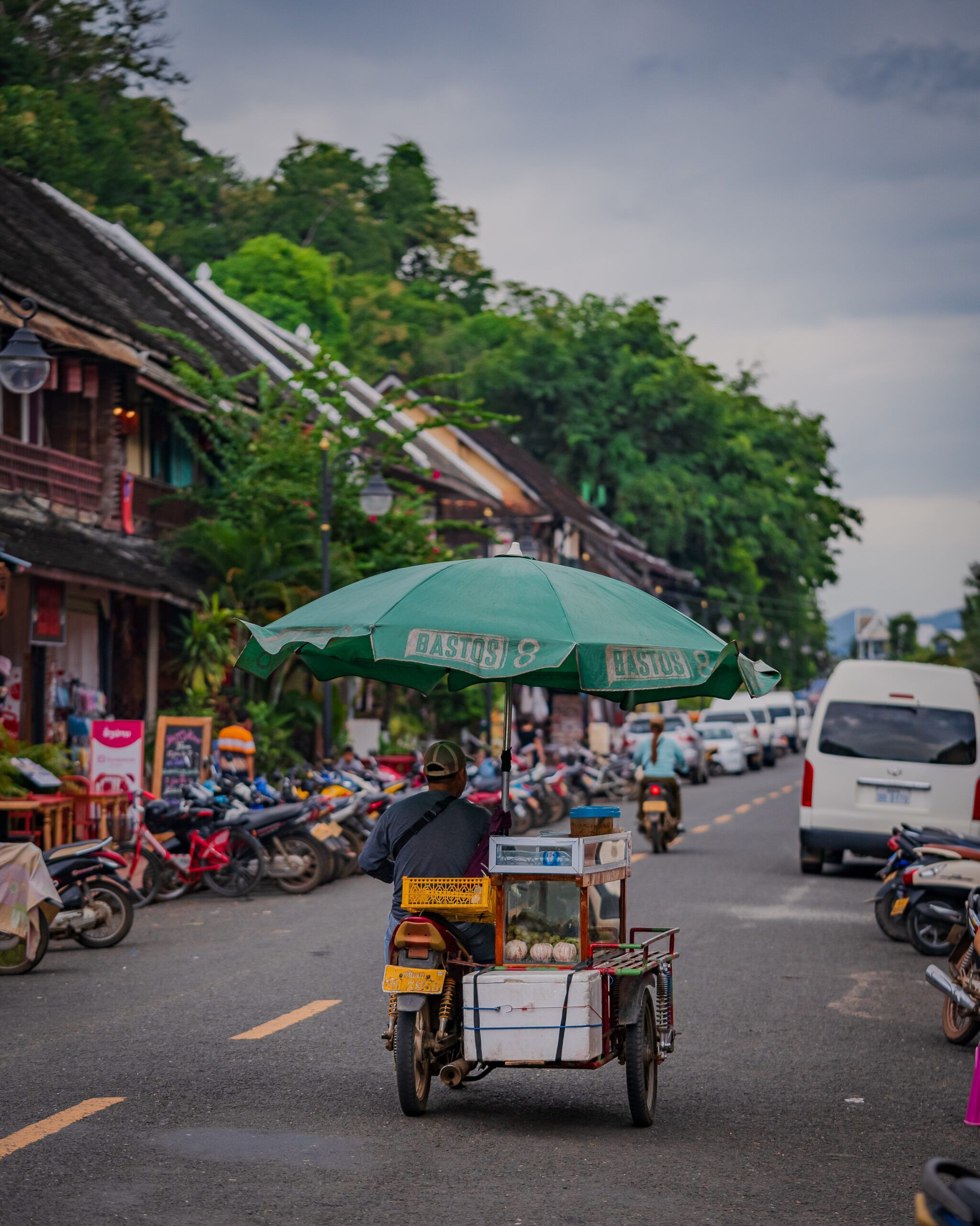 Mężczyzna na motorowerze z przyczepką i zielonym parasolem sprzedający jedzenie na ulicach Luang Prabang.
