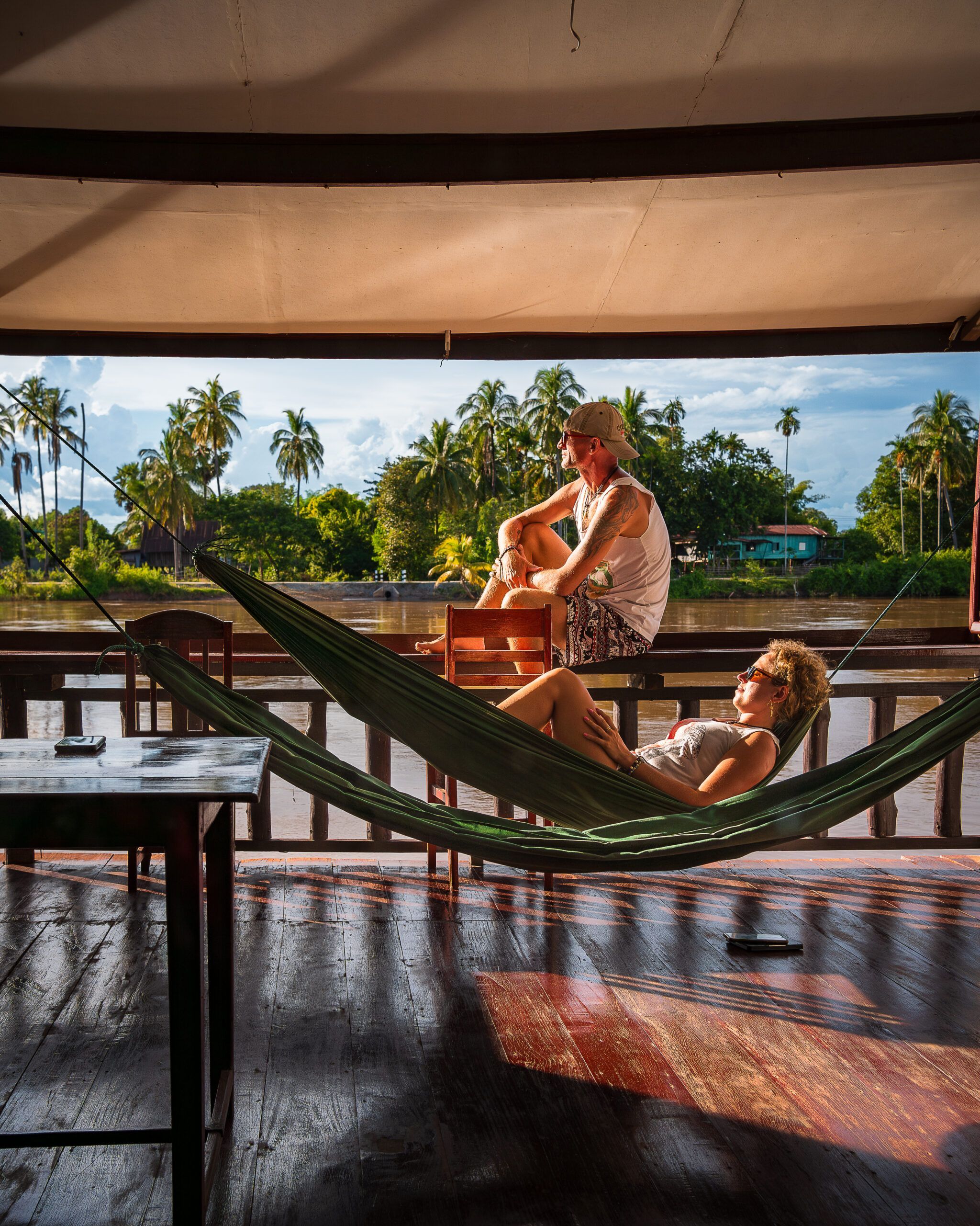 Para odpoczywa na tarasie nad rzeką w Laosie – mężczyzna siedzi na krześle, a kobieta relaksuje się w hamaku, w tle palmy kokosowe.
