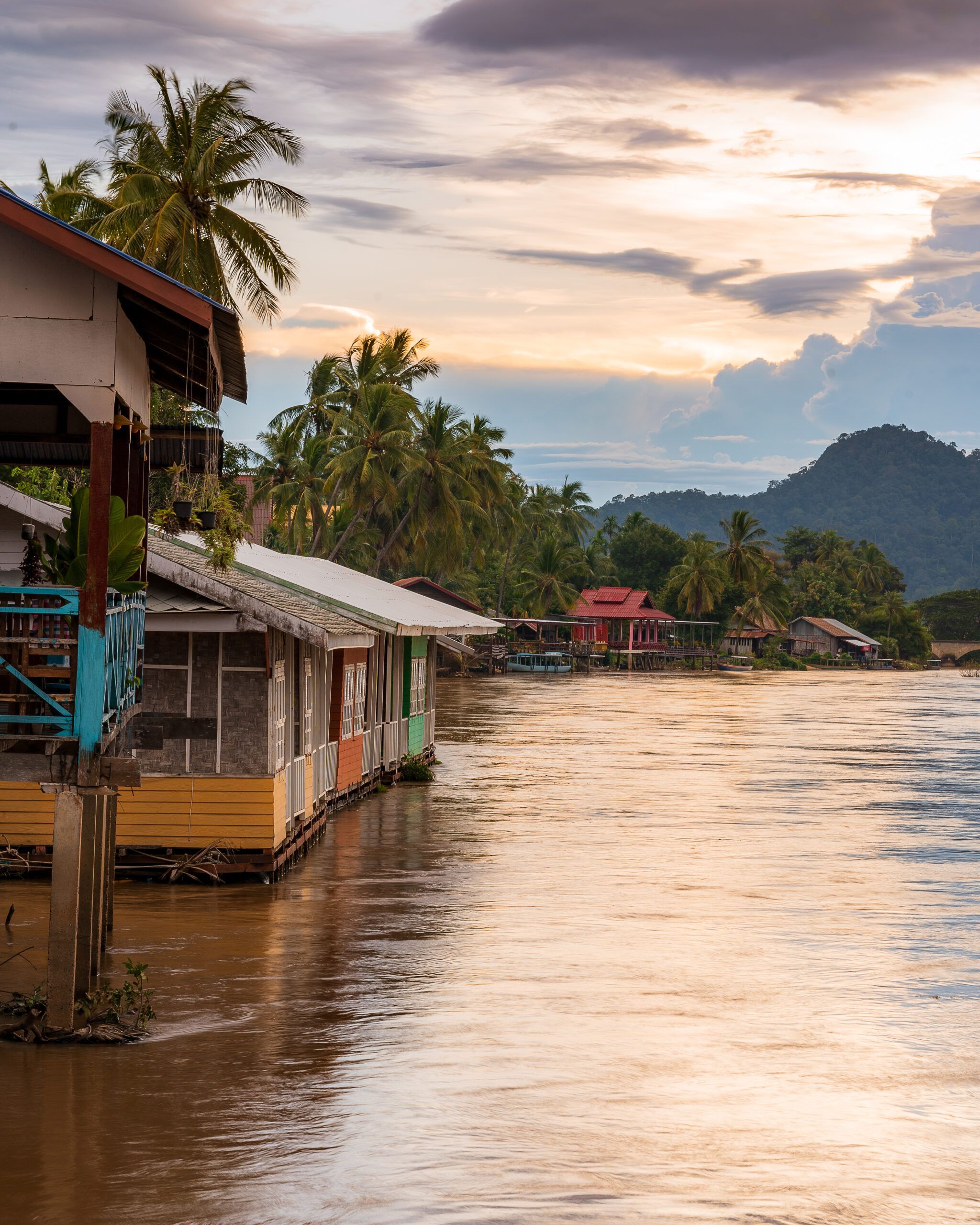 Kolorowe domy na palach nad brzegiem rzeki Mekong w Laosie, o zachodzie słońca, w otoczeniu palm i gór.
