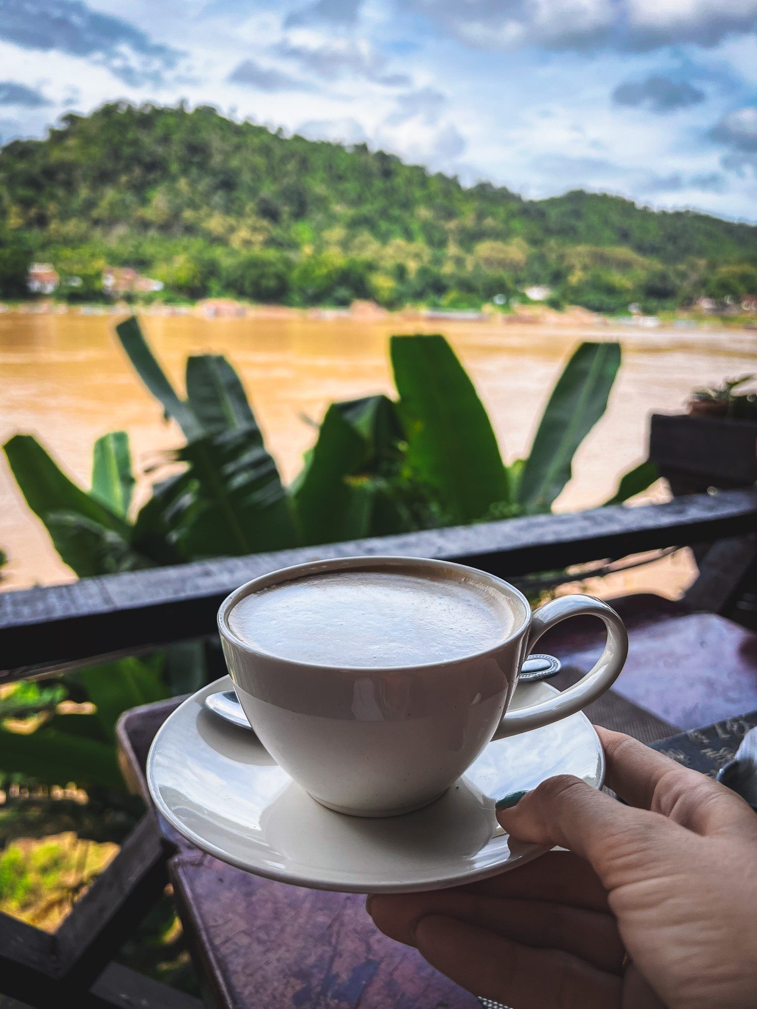Biała filiżanka cappuccino trzymana w dłoni z widokiem na rzekę Mekong i zielone wzgórza w tle.