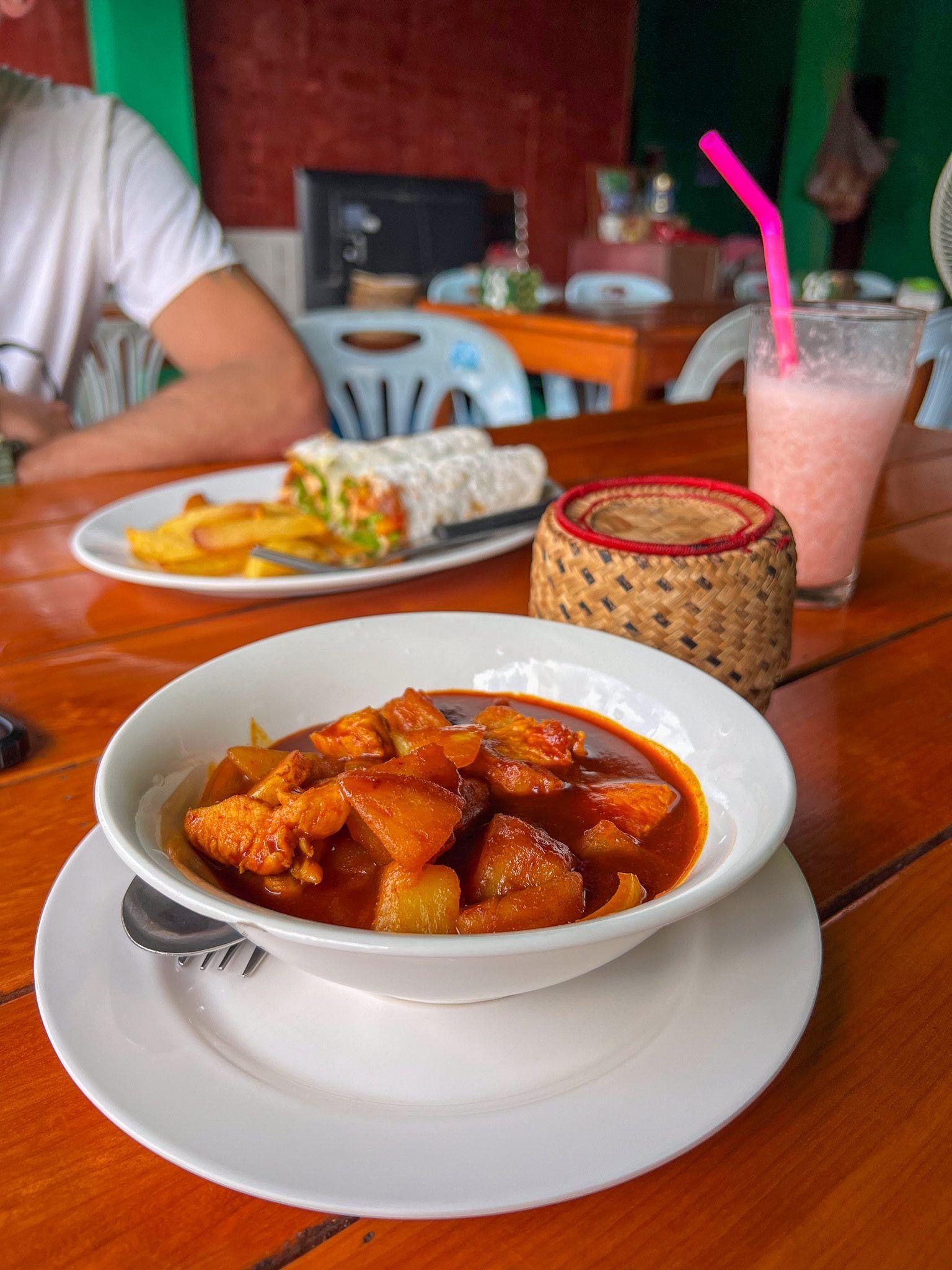 Talerz aromatycznego curry z kawałkami kurczaka i ziemniakami w gęstym, czerwonym sosie, podany w białej misce na drewnianym stole, w tle koktajl i inne danie.