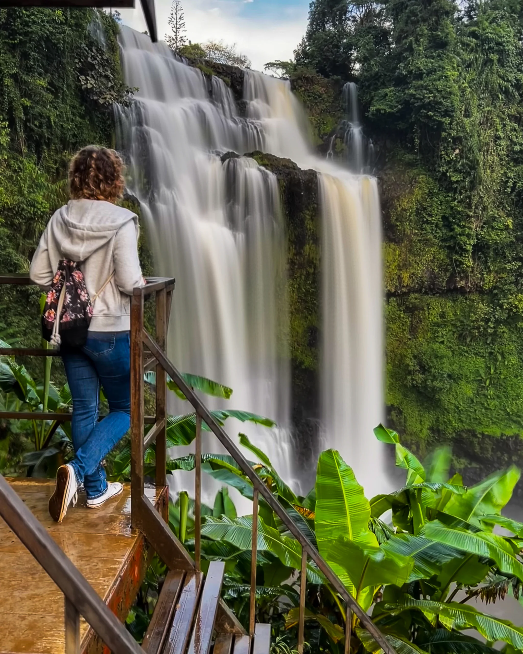 Kobieta stojąca na platformie widokowej, podziwiająca wysoki, wielopoziomowy wodospad otoczony tropikalną roślinnością.