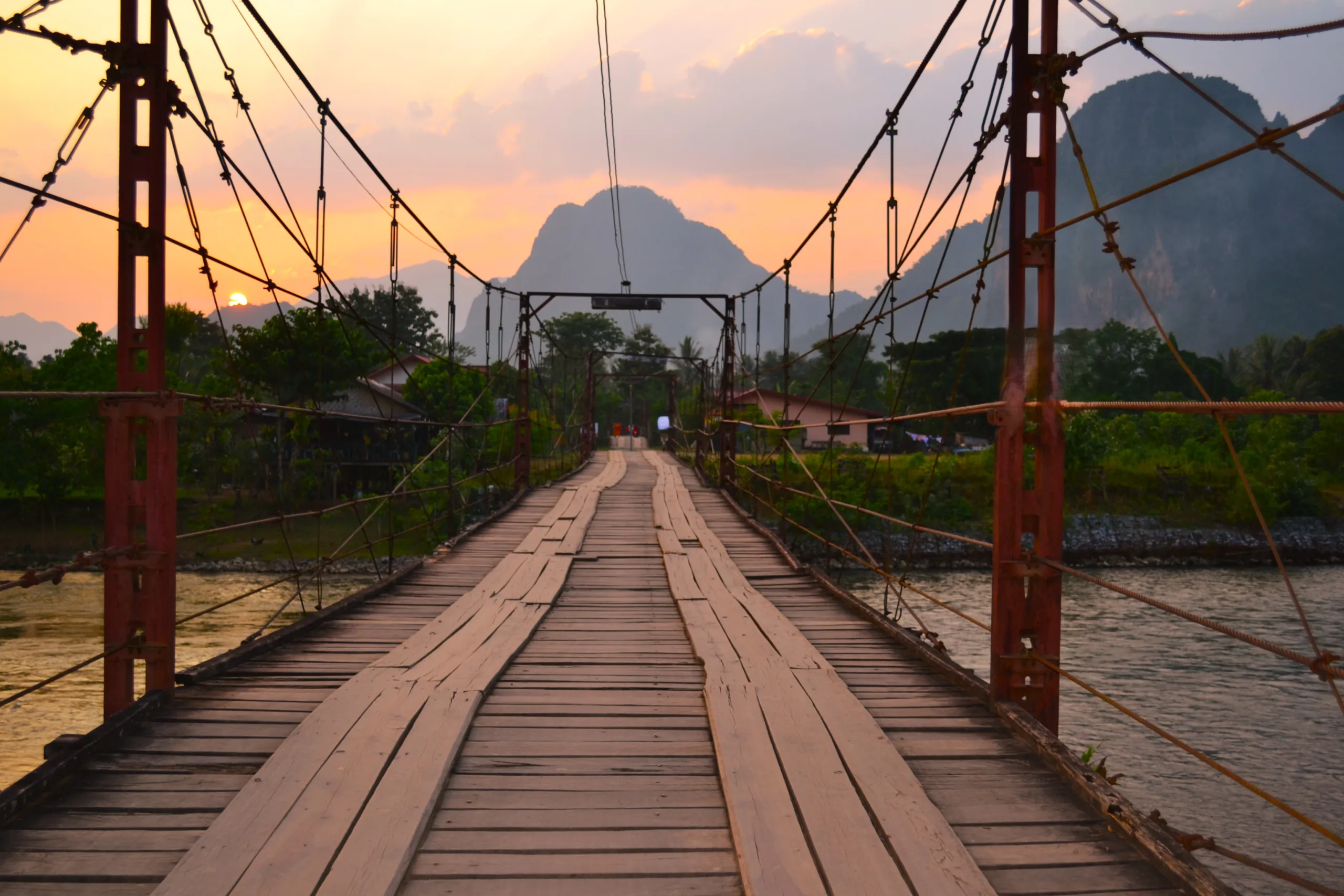 Stary drewniany most wiszący nad rzeką Nam Song w Laosie, prowadzący w stronę gór widocznych w tle podczas zachodu słońca.