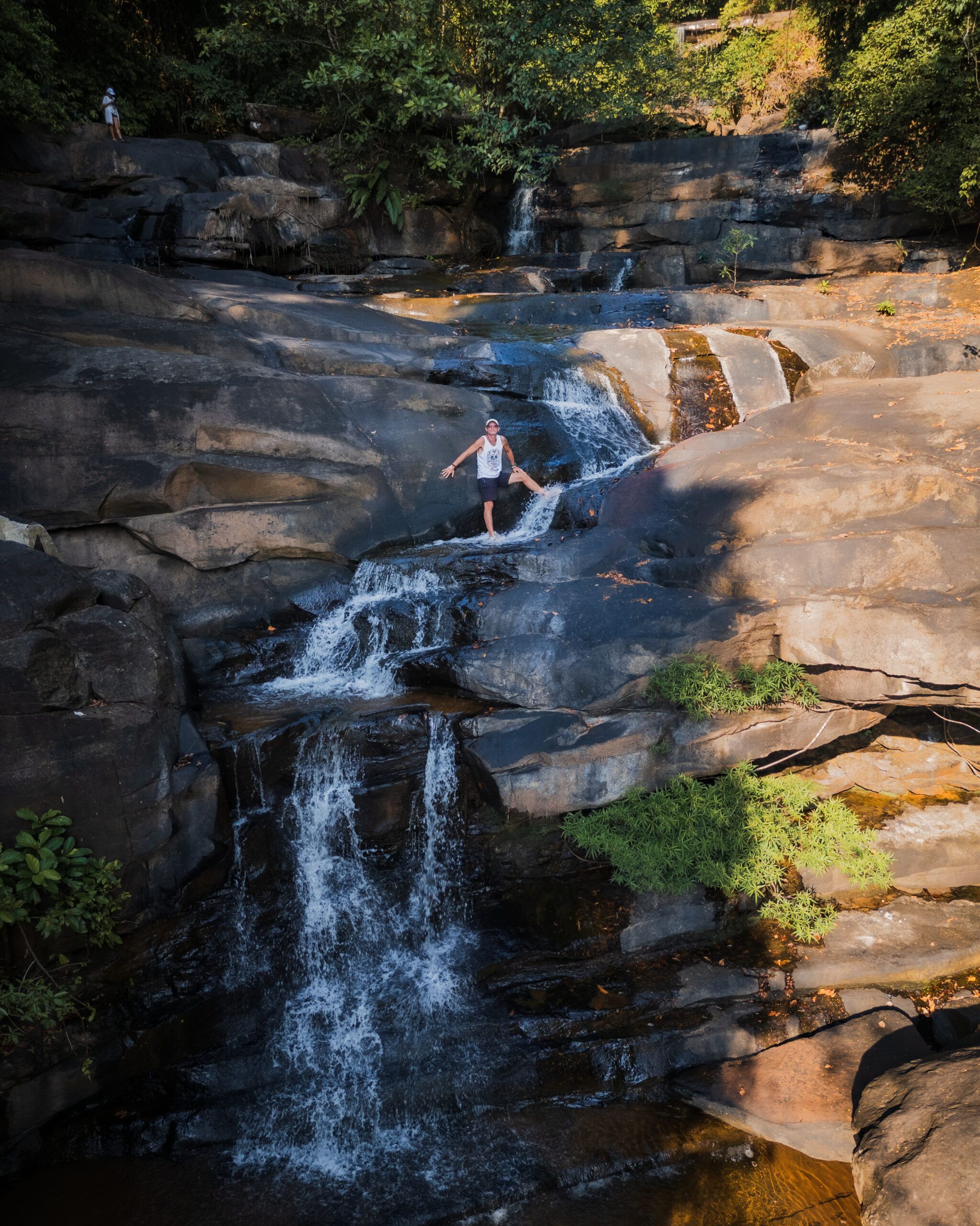 Osoba stojąca na naturalnych progach wodospadu wśród skał i tropikalnej roślinności
