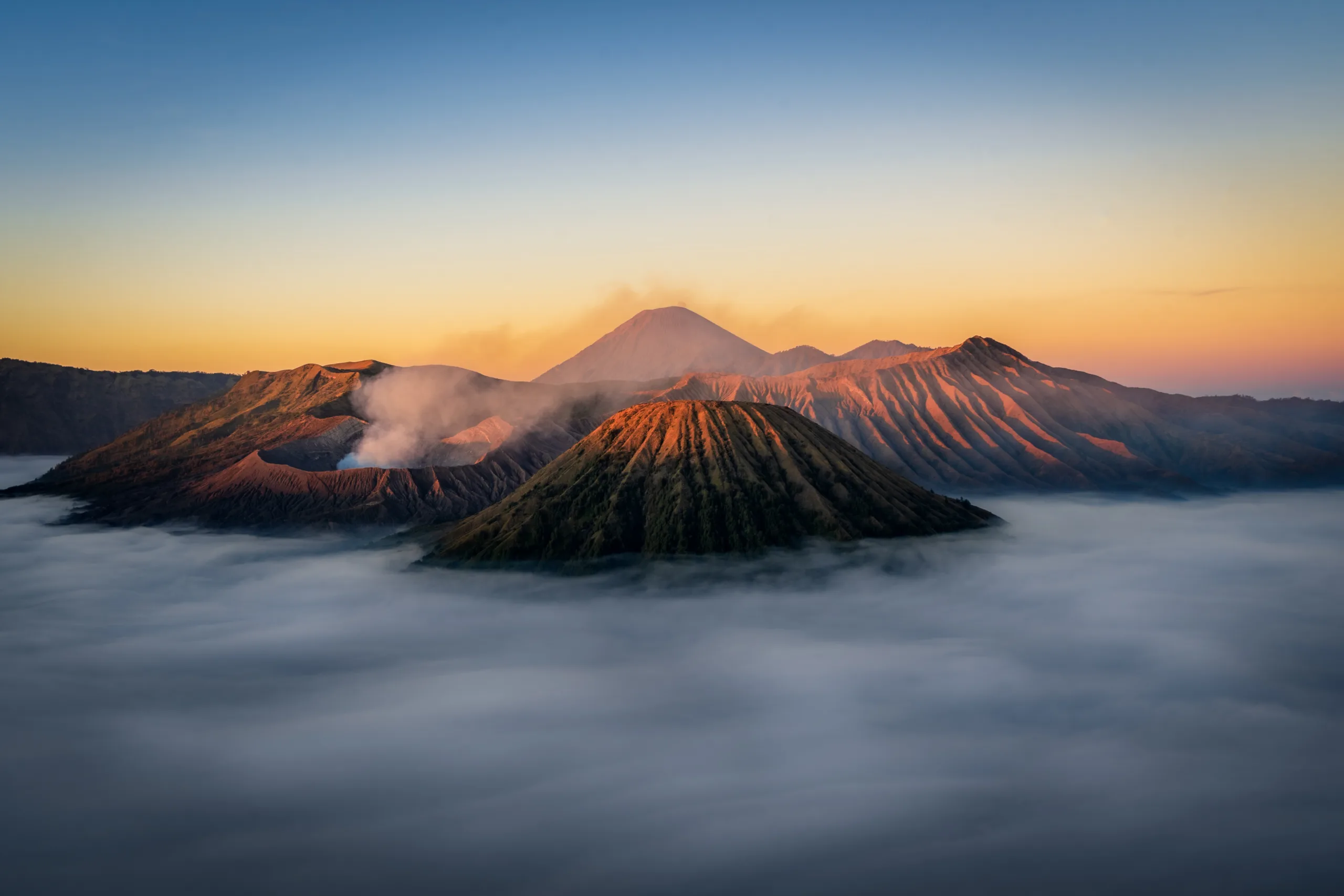 Panorama wulkanu Bromo o wschodzie słońca – krater z dymem unoszącym się nad krawędzią, otoczony górskimi szczytami i morzem chmur w Parku Narodowym Bromo Tengger Semeru na Jawie.