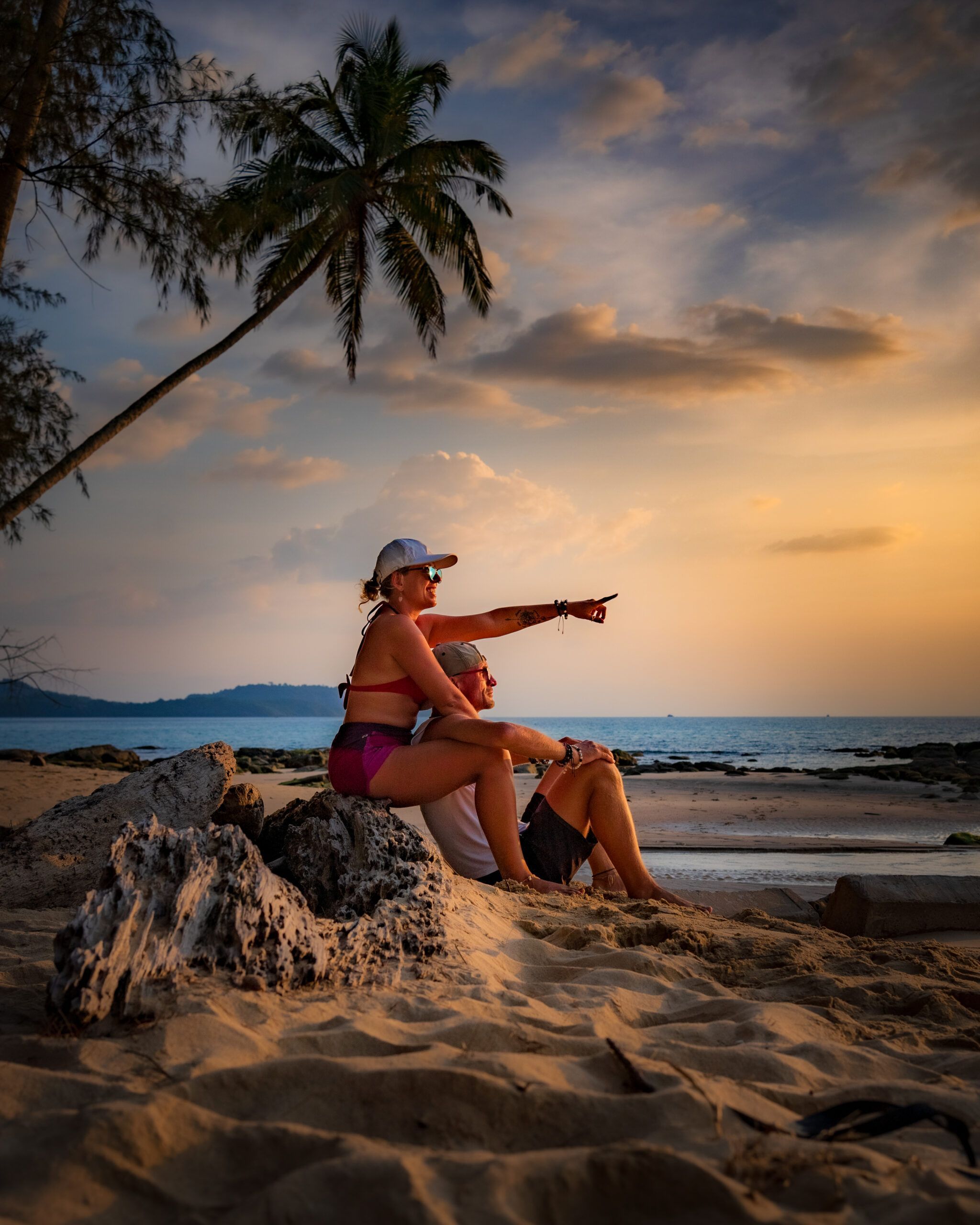 Para siedząca na plaży podczas zachodu słońca, kobieta pokazuje coś na horyzoncie