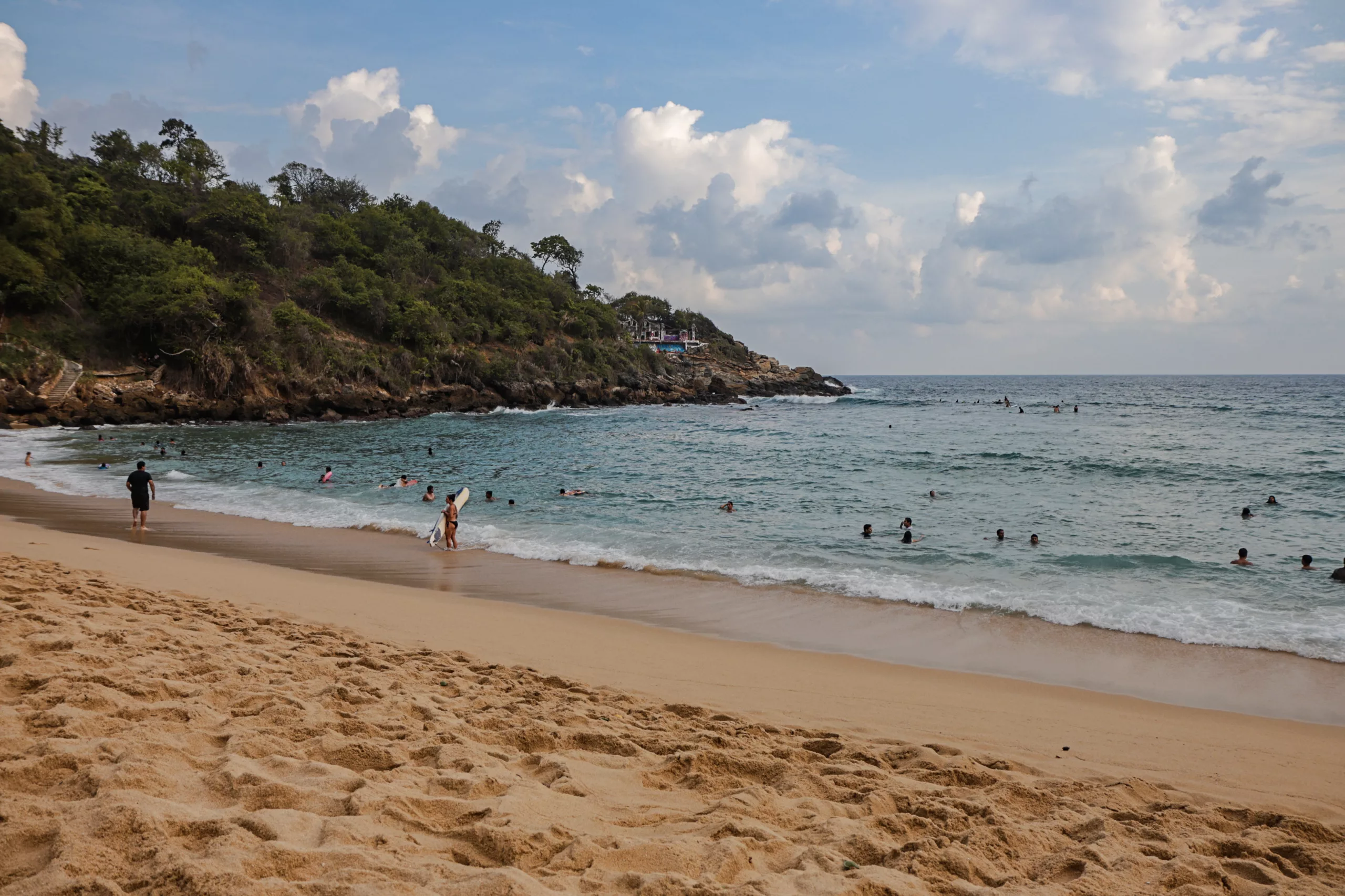 Plaża z delikatnym piaskiem i kąpiącymi się ludźmi, surfer trzyma deskę przy brzegu. W tle zielone wzgórze i skalisty cypel