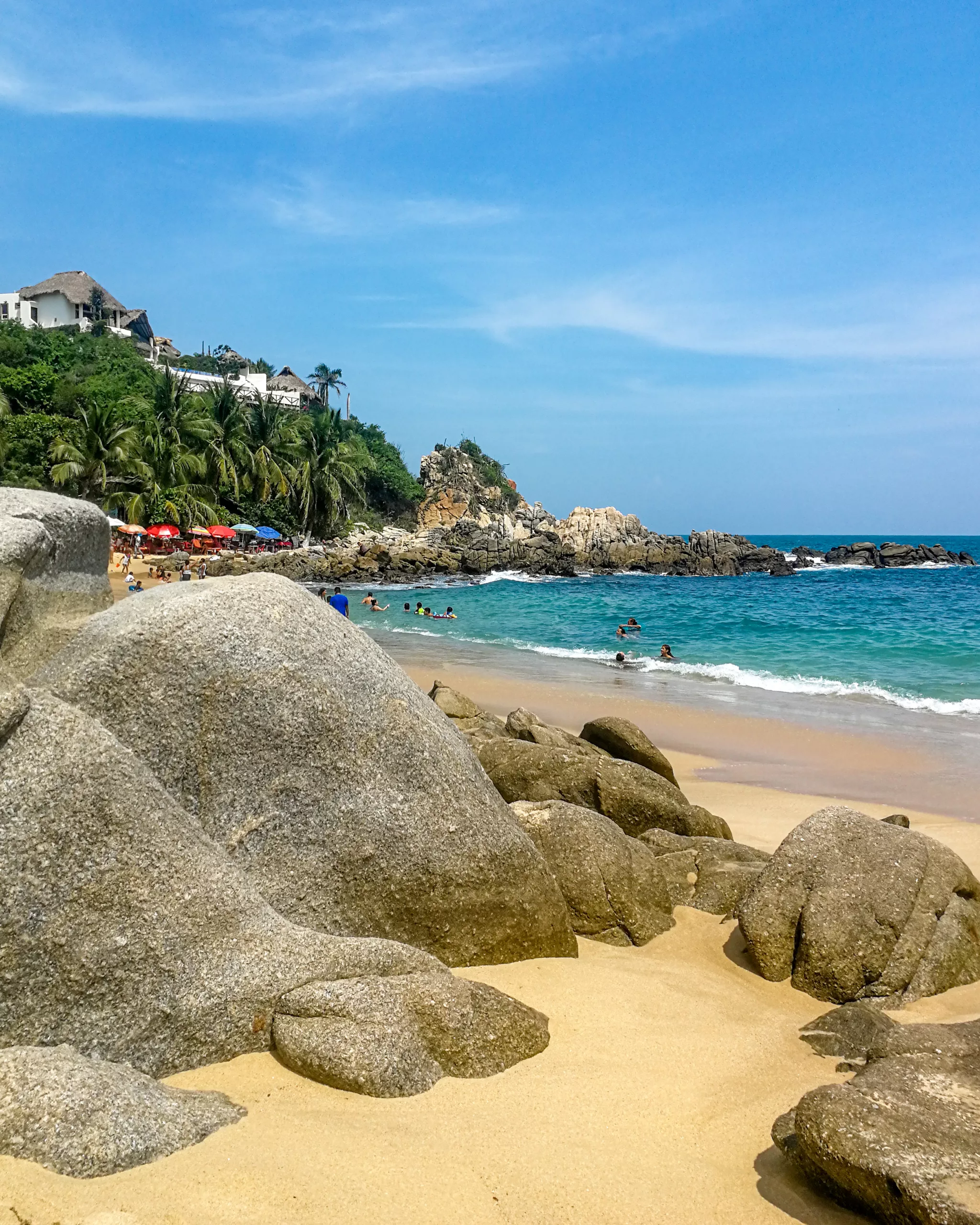 Widok na kamienistą plażę z dużymi głazami na pierwszym planie, ludzie kąpią się w turkusowej wodzie, a w tle widoczne są palmy i zabudowania na wzgórzu