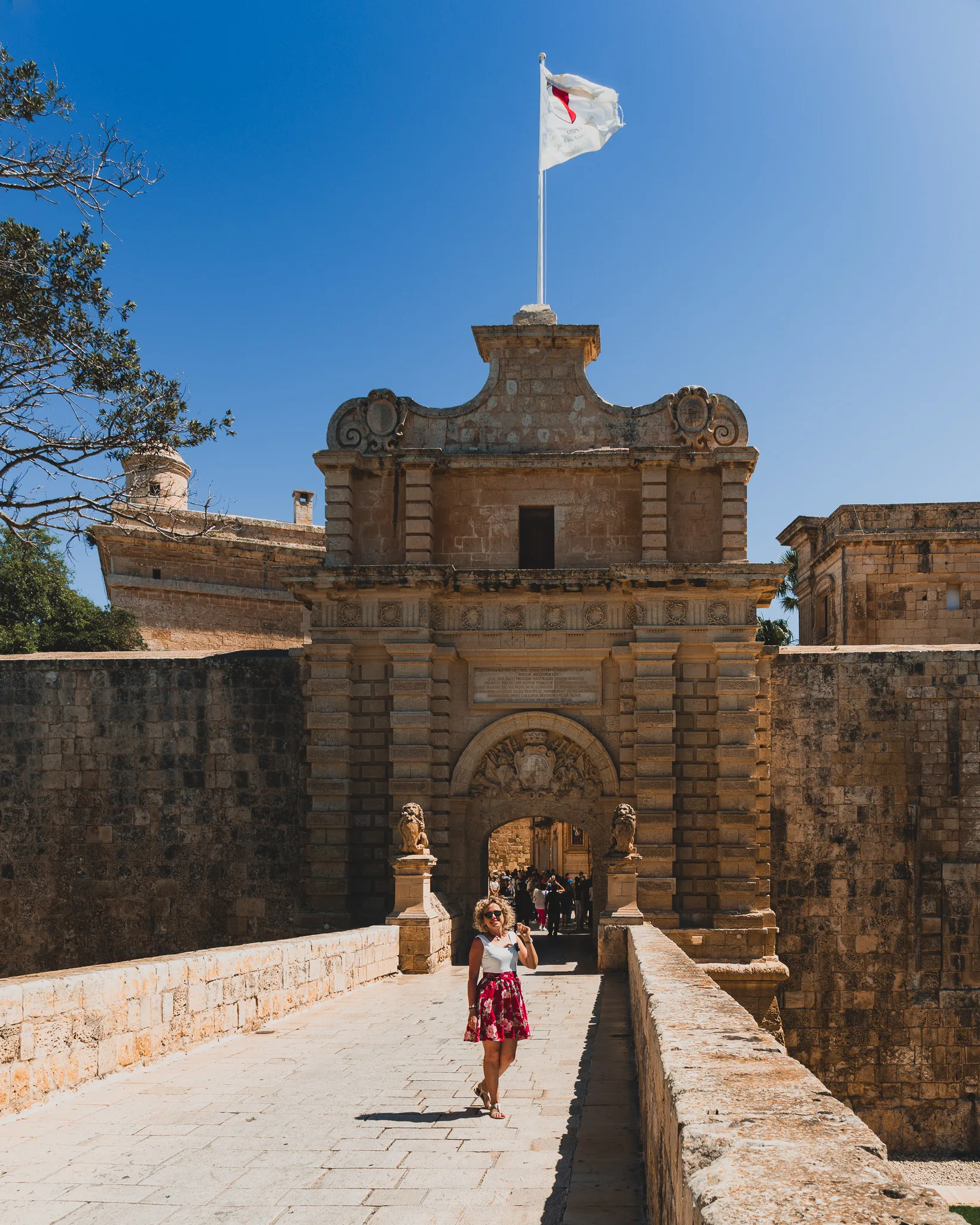 Brama Mdina Gate na Malcie z kobietą w czerwonej spódnicy spacerującą po kamiennym moście pod błękitnym niebem