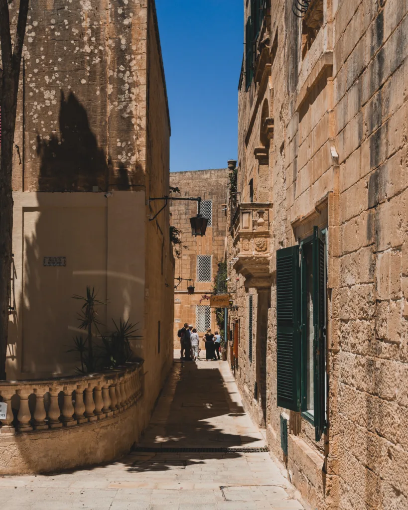 Wąska uliczka w Mdinie na Malcie z grupą turystów i kamiennymi budynkami w cieniu południowego słońca