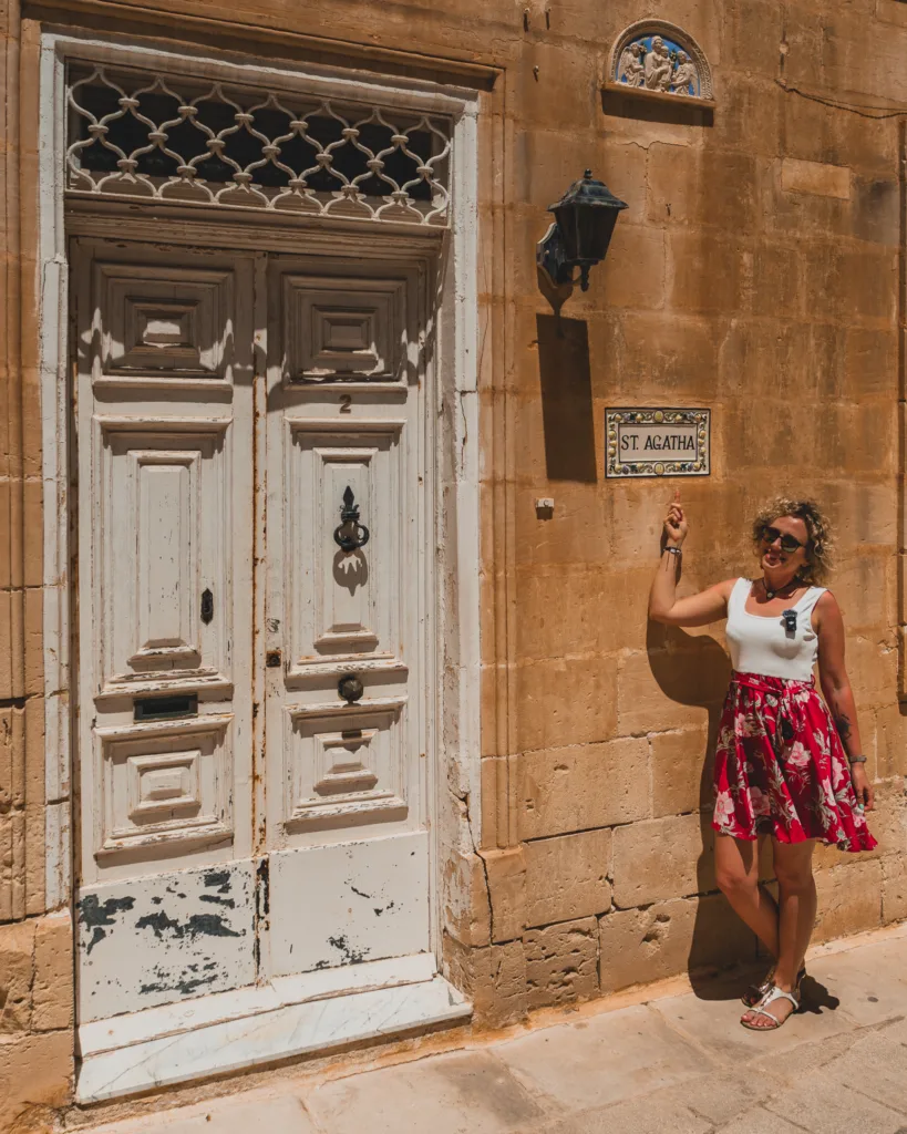 Turystka wskazująca ceramiczną tabliczkę z napisem 'St. Agatha' na tle piaskowego budynku z białymi drzwiami w Mdinie na Malcie