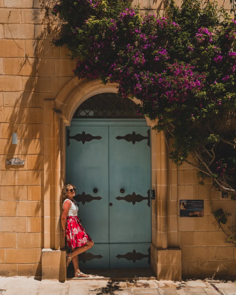 Kobieta w czerwonej spódnicy oparta o pastelowe, błękitne drzwi budynku na Malcie, ozdobionego fioletowym bugenwillą