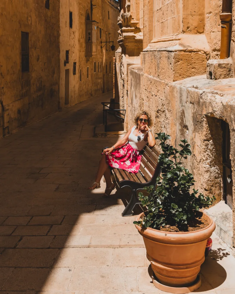 Kobieta w czerwonej spódnicy siedzi na ławce w wąskiej, zacienionej uliczce Mdiny na Malcie