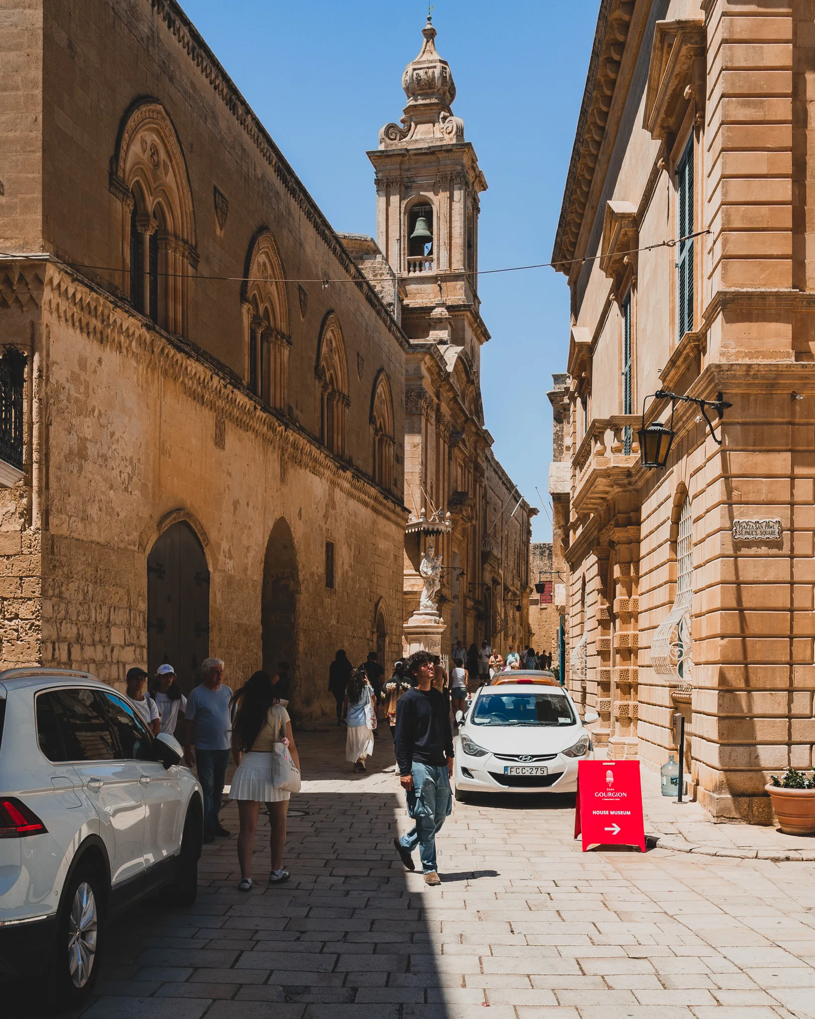 Ulica w Mdinie z zabytkowymi budynkami, dzwonnicą i turystami spacerującymi w słoneczny dzień
