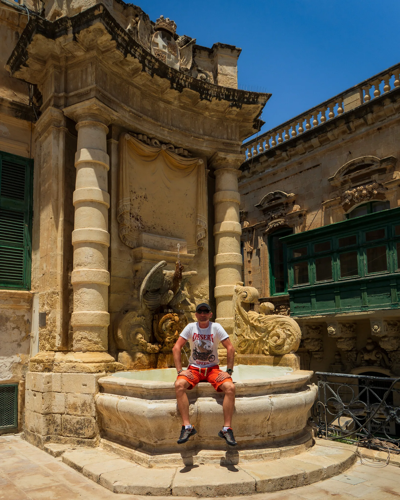 Mężczyzna siedzący na zabytkowej fontannie z rzeźbą ryby przy barokowej fasadzie budynku – Valletta, Malta