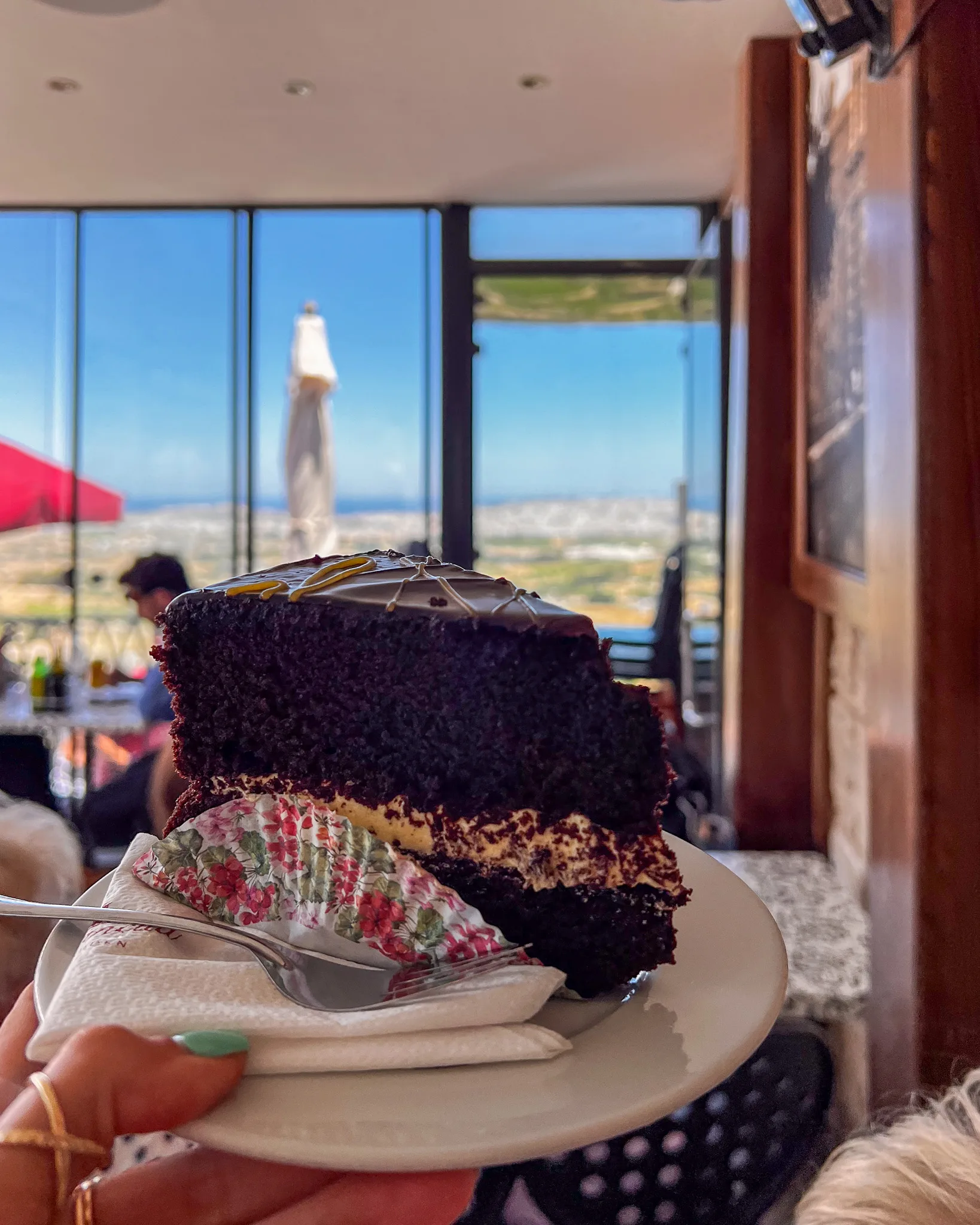 Kawałek tortu czekoladowego trzymany przed panoramicznym widokiem na Maltę z kawiarni Fontanella
