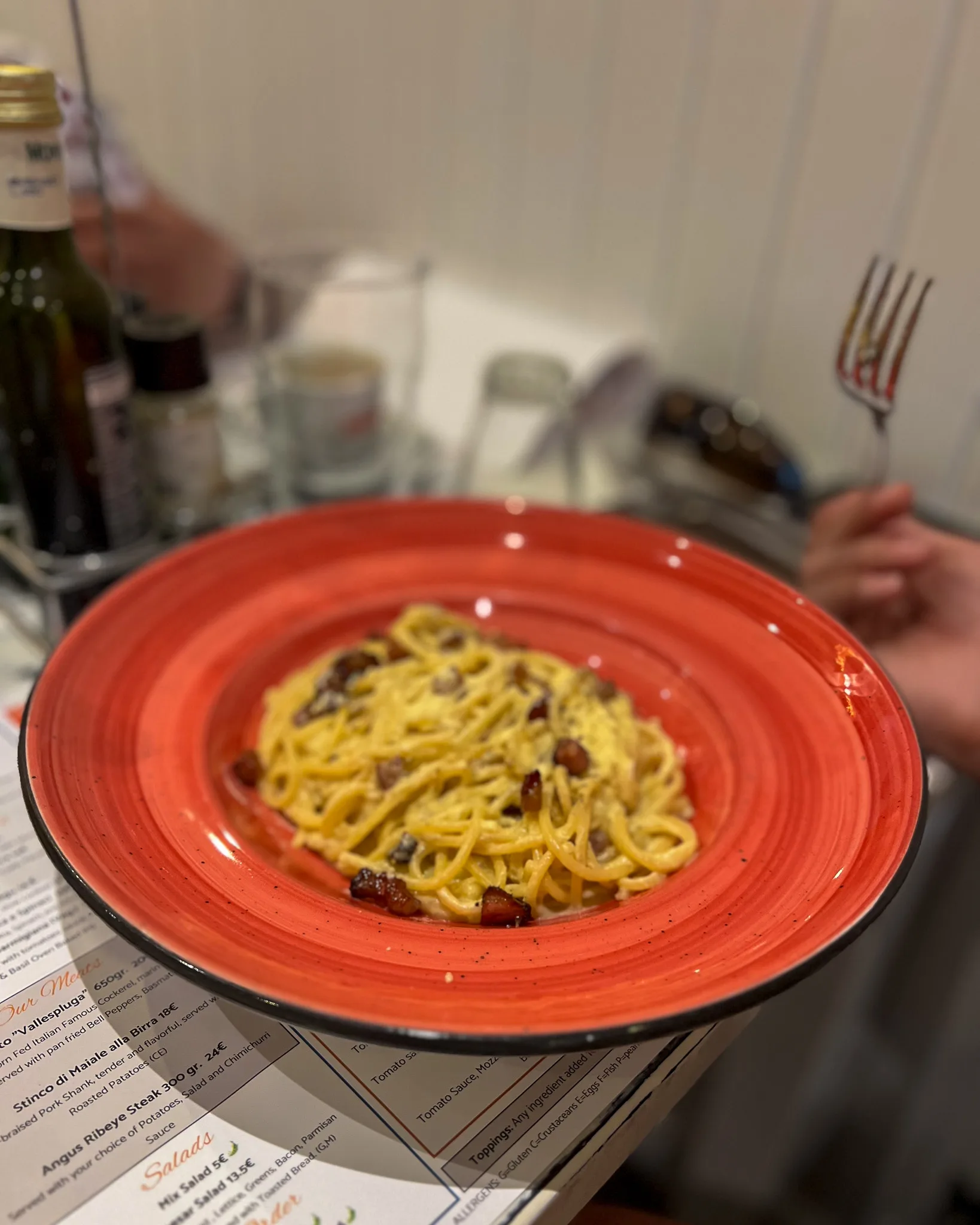 Porcja klasycznego spaghetti carbonara podana na czerwonym talerzu w restauracji, z widocznymi kawałkami boczku i posypką z sera