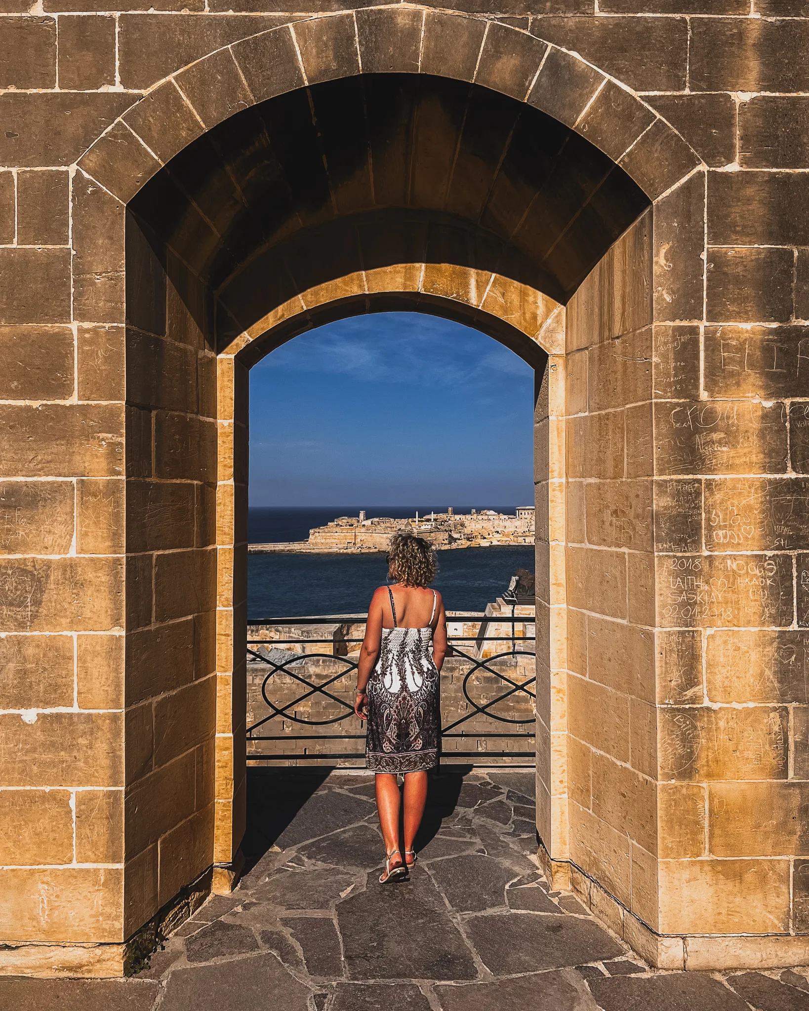 Kobieta w sukience patrzy przez łukowate przejście na morze i Fort Ricasoli – Valletta, Malta