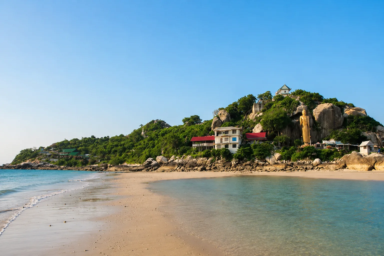 Spokojna plaża o poranku z jasnym piaskiem, błękitną wodą, zielonym wzgórzem i posągiem Buddy w tle