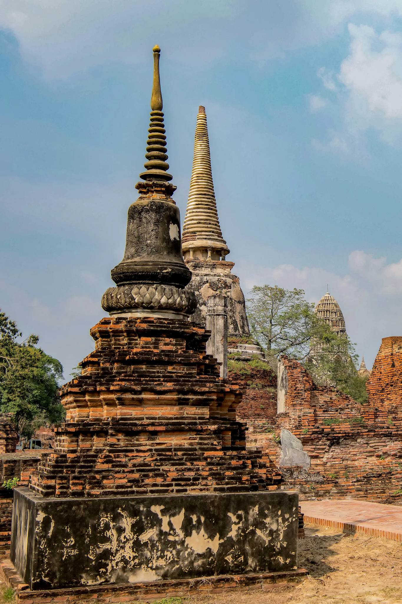 Starożytna ceglana prangowa wieża świątynna w Ayutthayi, częściowo porośnięta roślinnością i otoczona ruinami dawnego kompleksu.