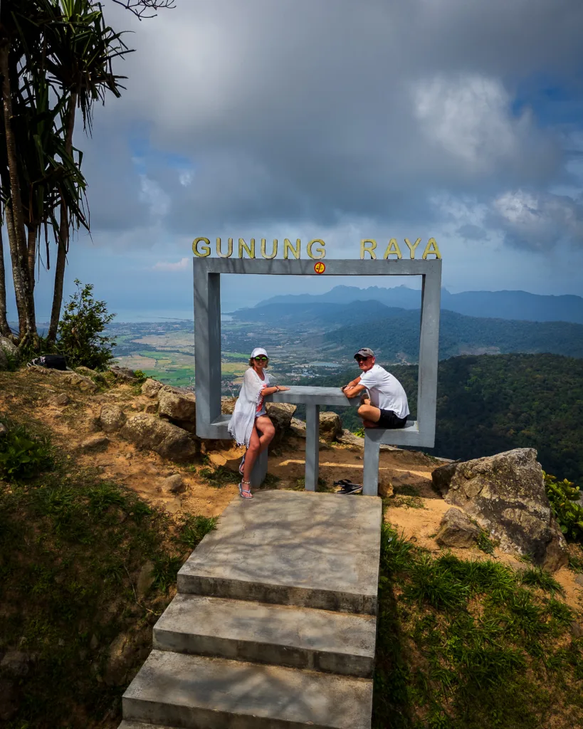 Para turystów siedząca w betonowej ramie widokowej na szczycie Gunung Raya na wyspie Langkawi w Malezji – panorama gór, dżungli i morza w tle