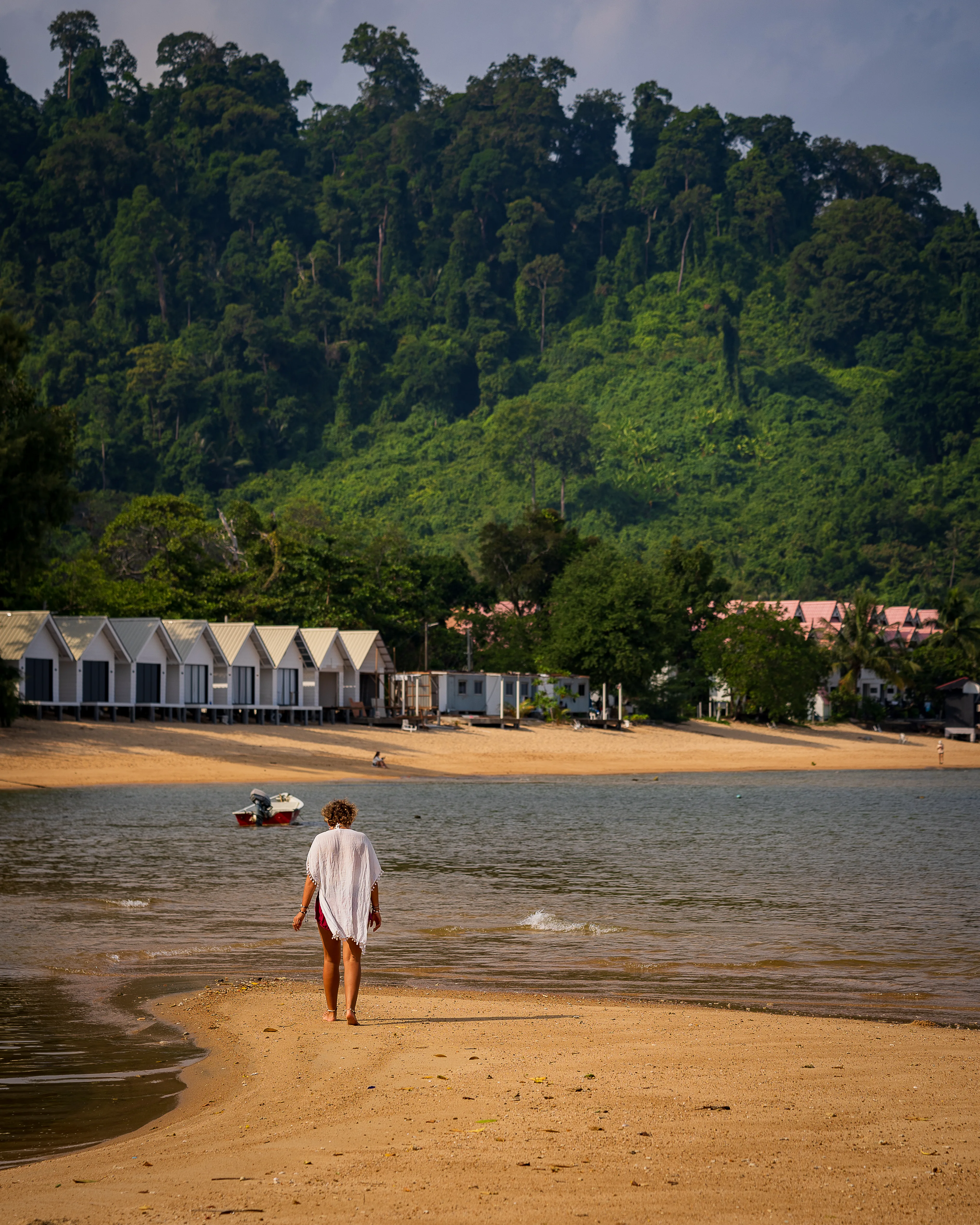 Podróżniczka spacerująca po spokojnej plaży na wyspie Tioman w Malezji, z tropikalnym lasem i bungalowami w tle.