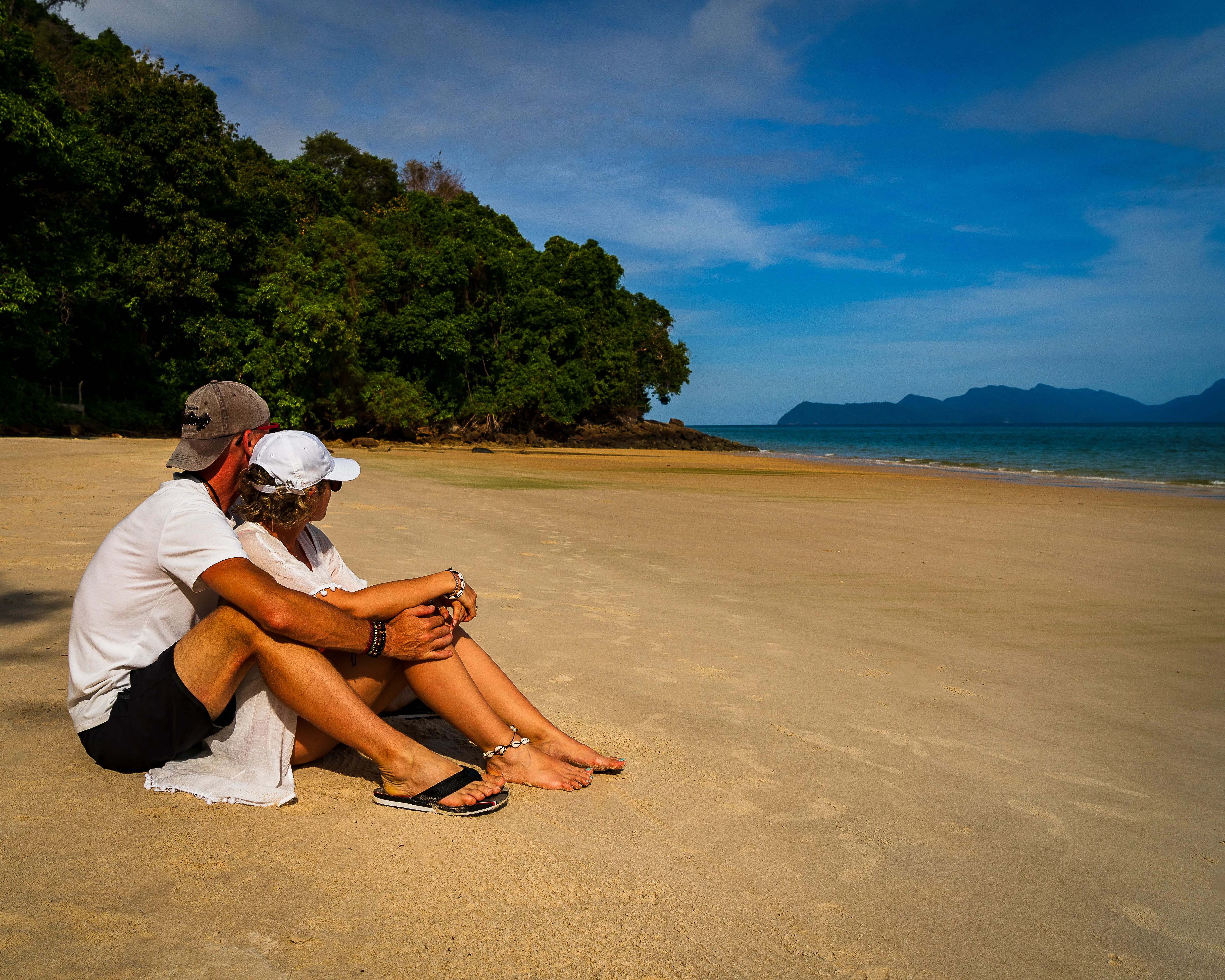 Para podróżników odpoczywająca na szerokiej, piaszczystej plaży na Langkawi w Malezji, z morzem i górzystymi wyspami na horyzoncie.