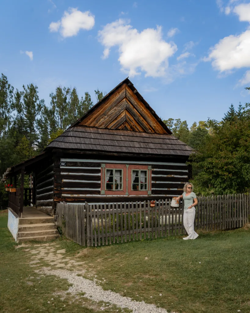Drewniana chatka w stylu ludowym z kolorowymi okiennicami, ogrodzona płotem, przed którą stoi kobieta - Skansen w Lublowej.