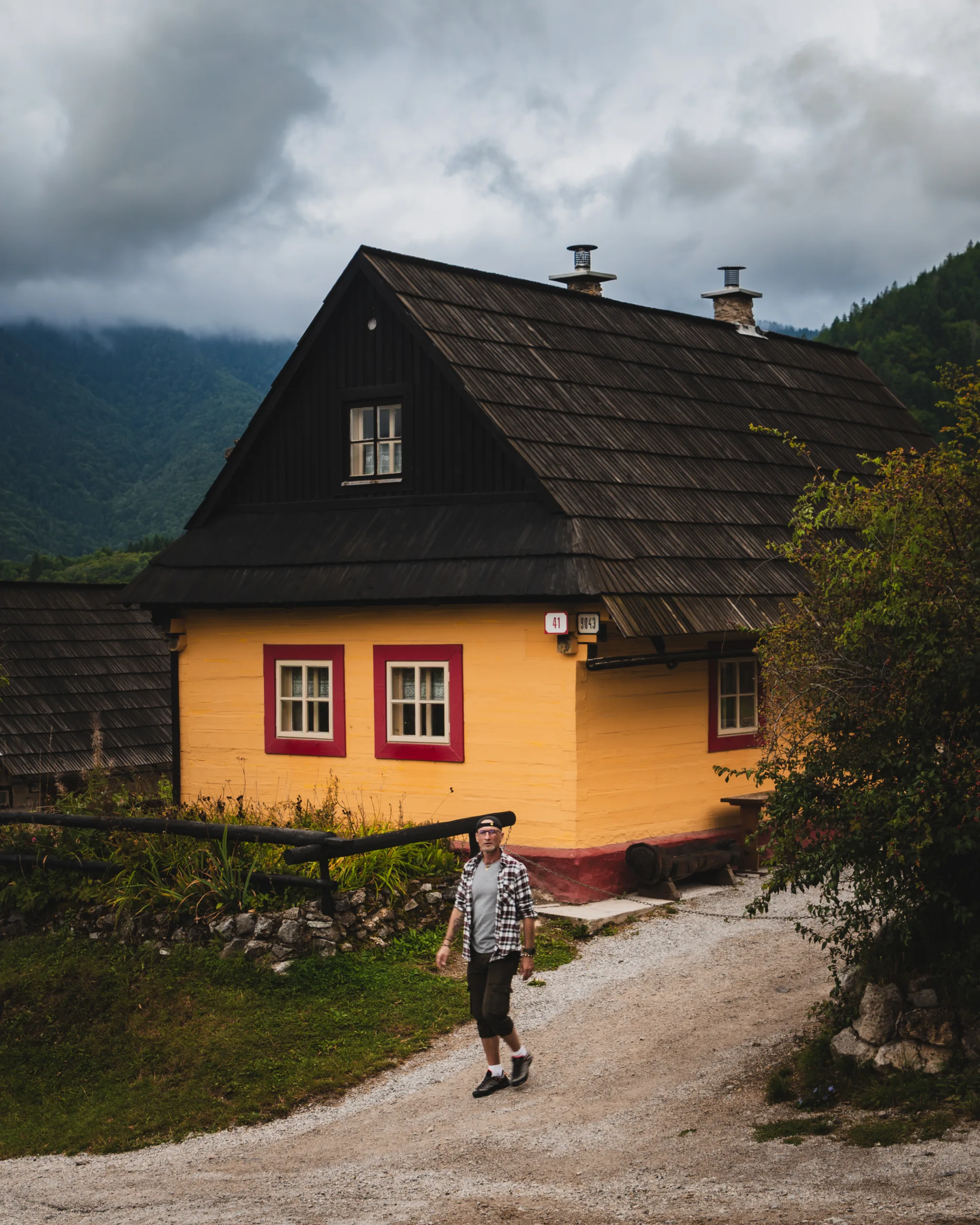 Tradycyjny drewniany dom we wsi Vlkolínec na Słowacji, pomalowany na żółto z czerwonymi ramami okien, wpisanej na listę UNESCO. Na pierwszym planie mężczyzna spacerujący po kamienistej drodze.