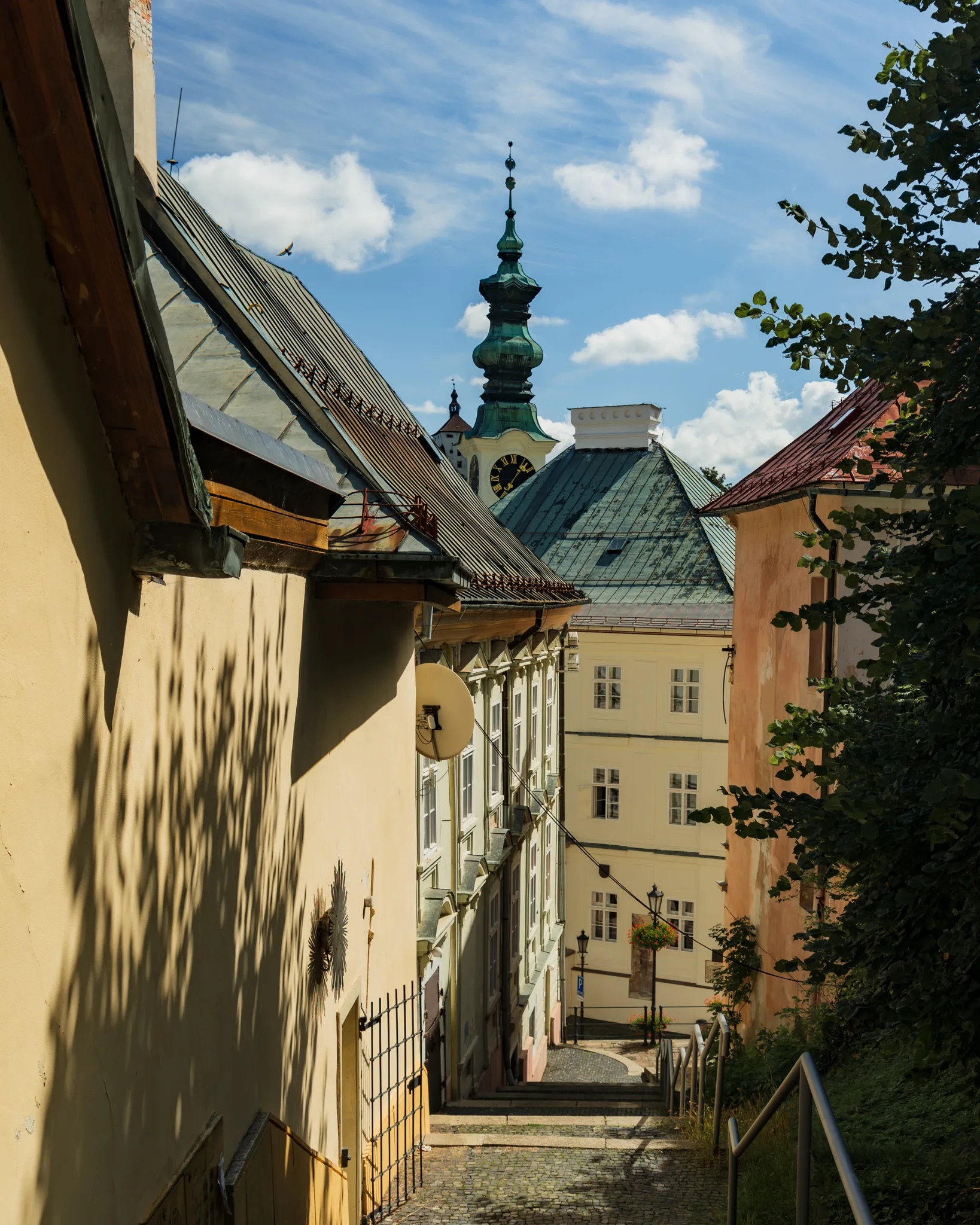 Wąska uliczka z brukowanymi schodami w Bańskiej Szczawnicy, prowadząca w dół pomiędzy kamienicami z widokiem na wieżę zegarową kościoła św. Katarzyny.