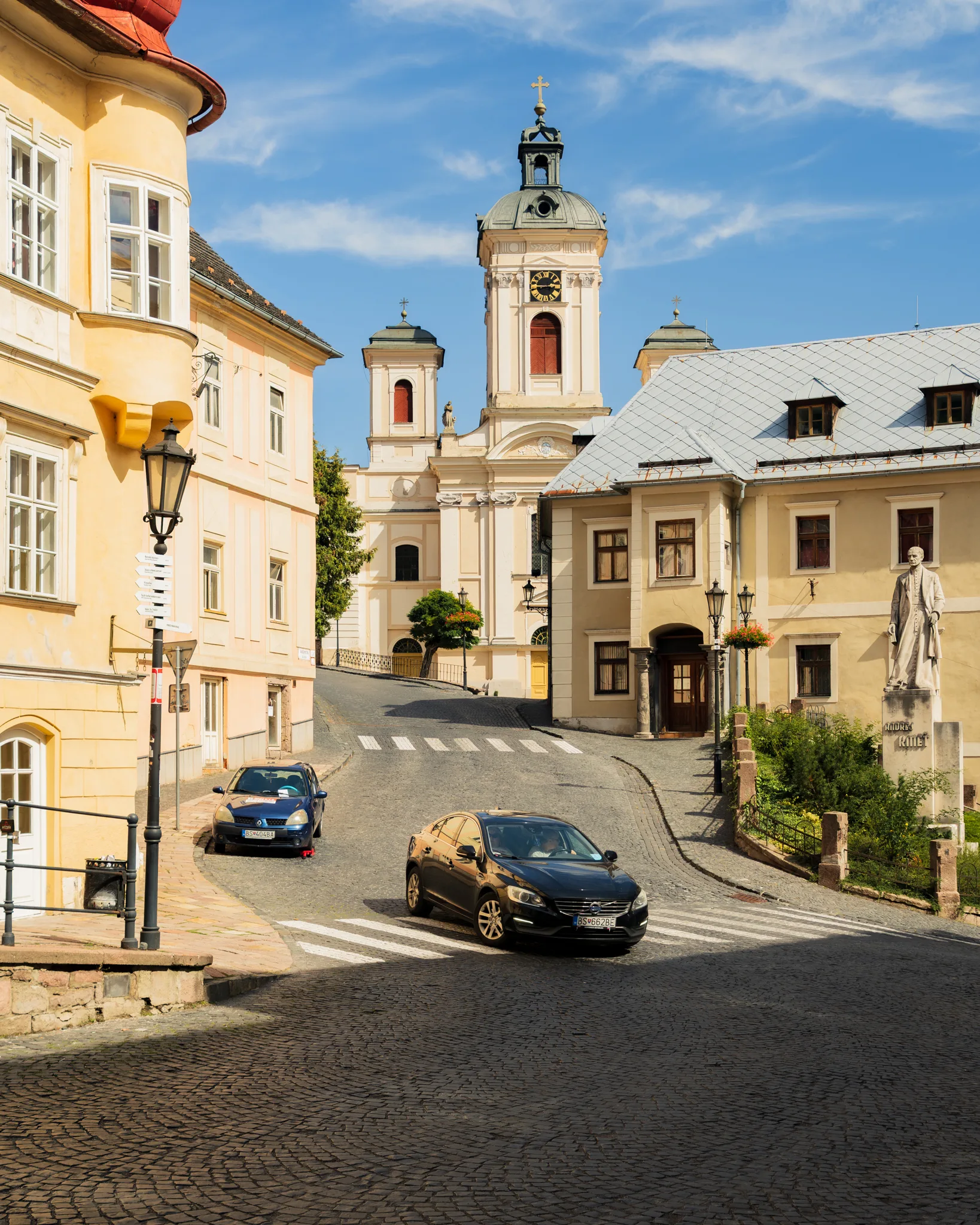 Widok na barokowy kościół św. Katarzyny w Bańska Szczawnica z perspektywy brukowanej uliczki z samochodami i zabytkowymi kamienicami po bokach.